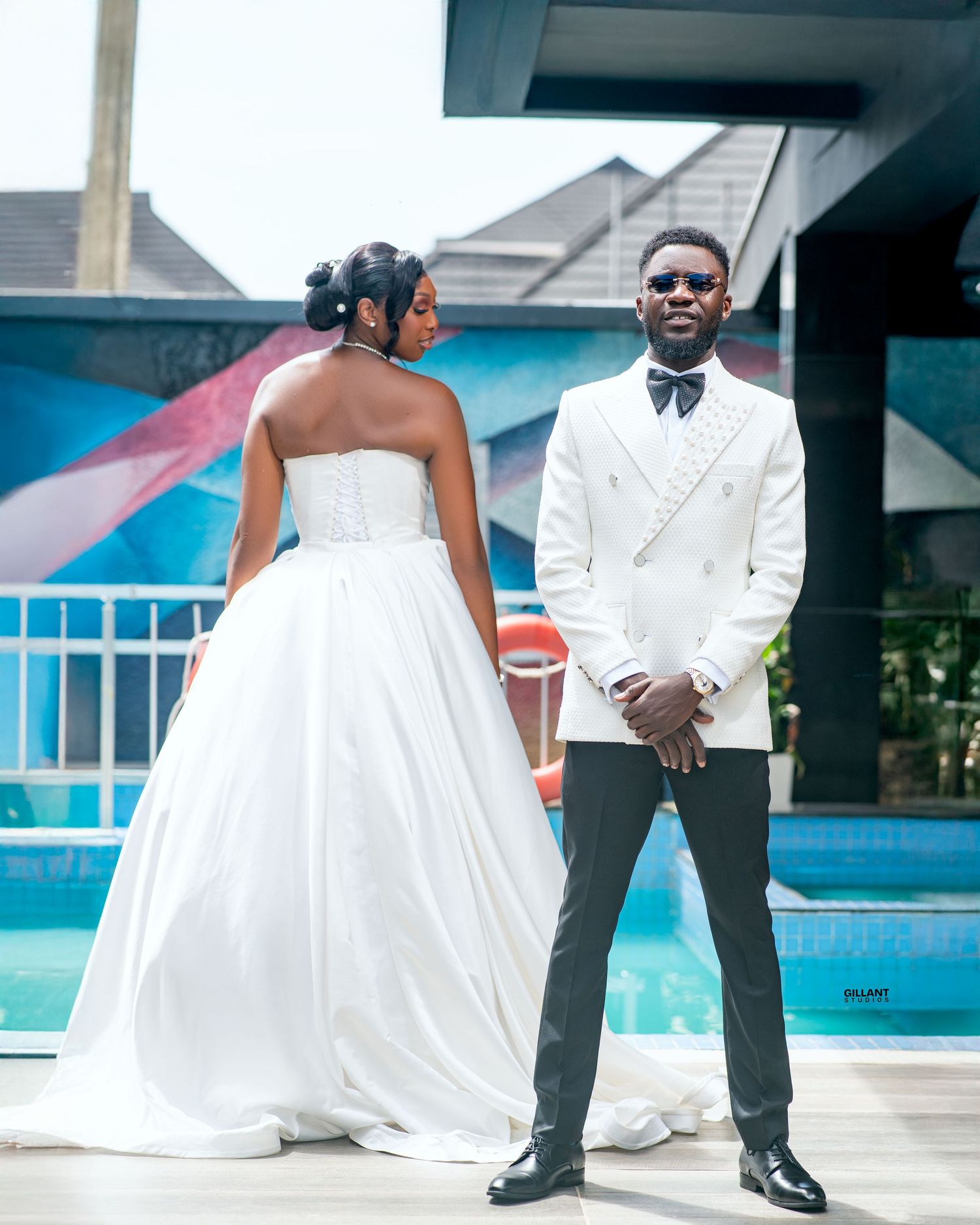 Wedding couple in white dress and tuxedo posing by pool with blue exterior.