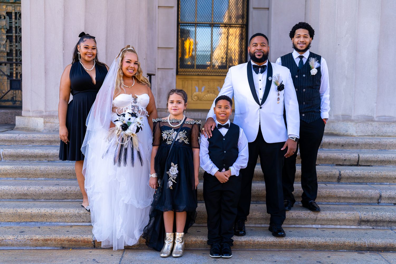 A formal wedding party stands on stone steps in front of pillars with a bride in white holding a bouquet.