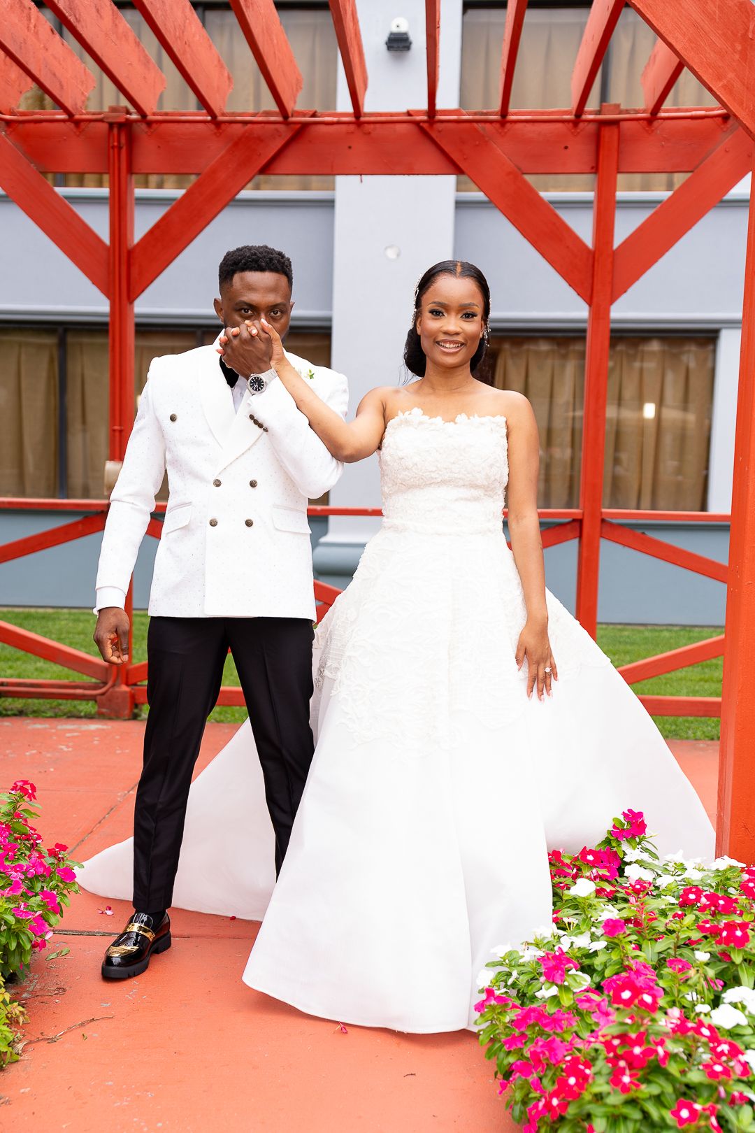 Romantic wedding photoshoot with couple in white attire against red architectural backdrop with pink flowers.