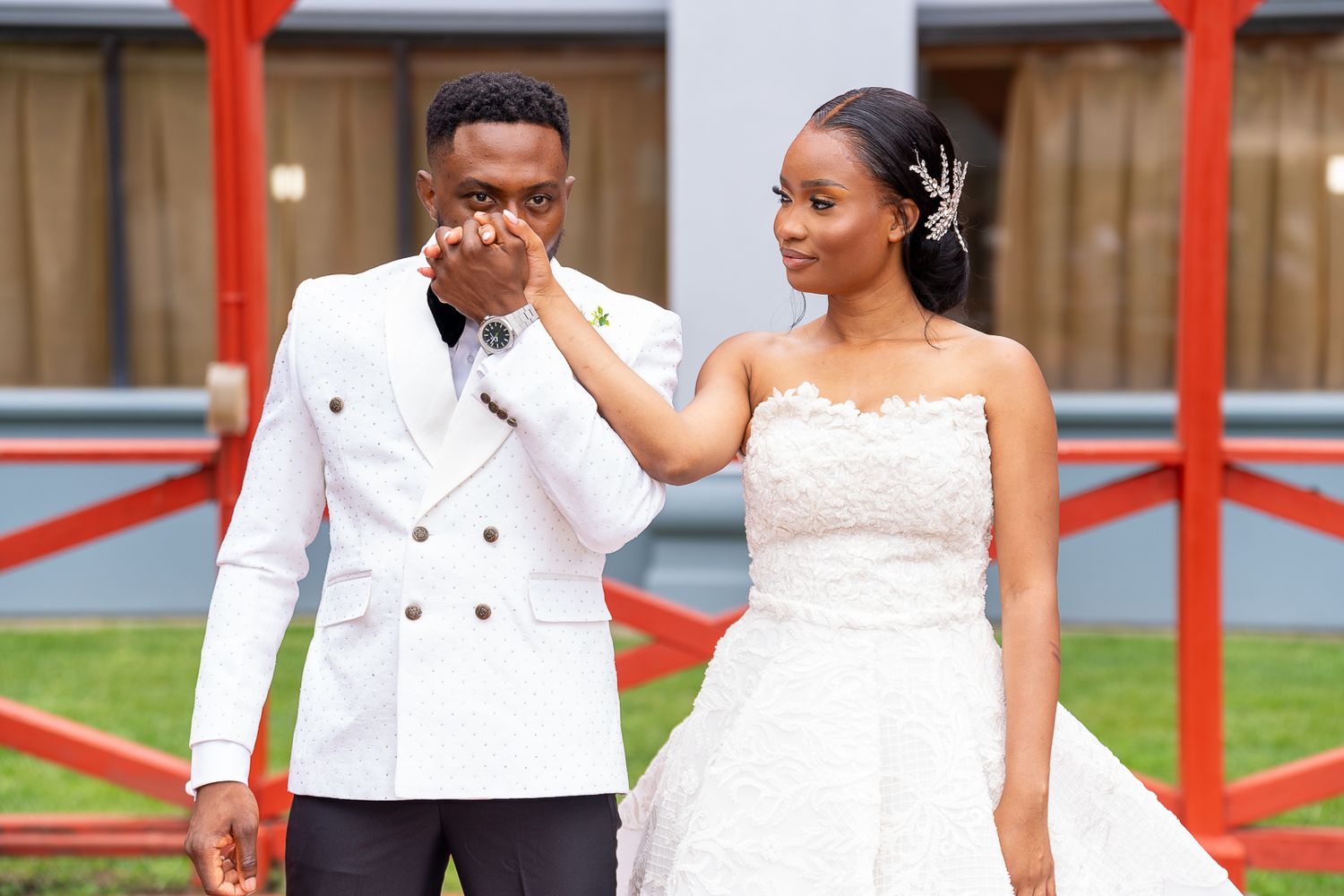 Tender moment between newlyweds in white formal attire on red-accented porch.