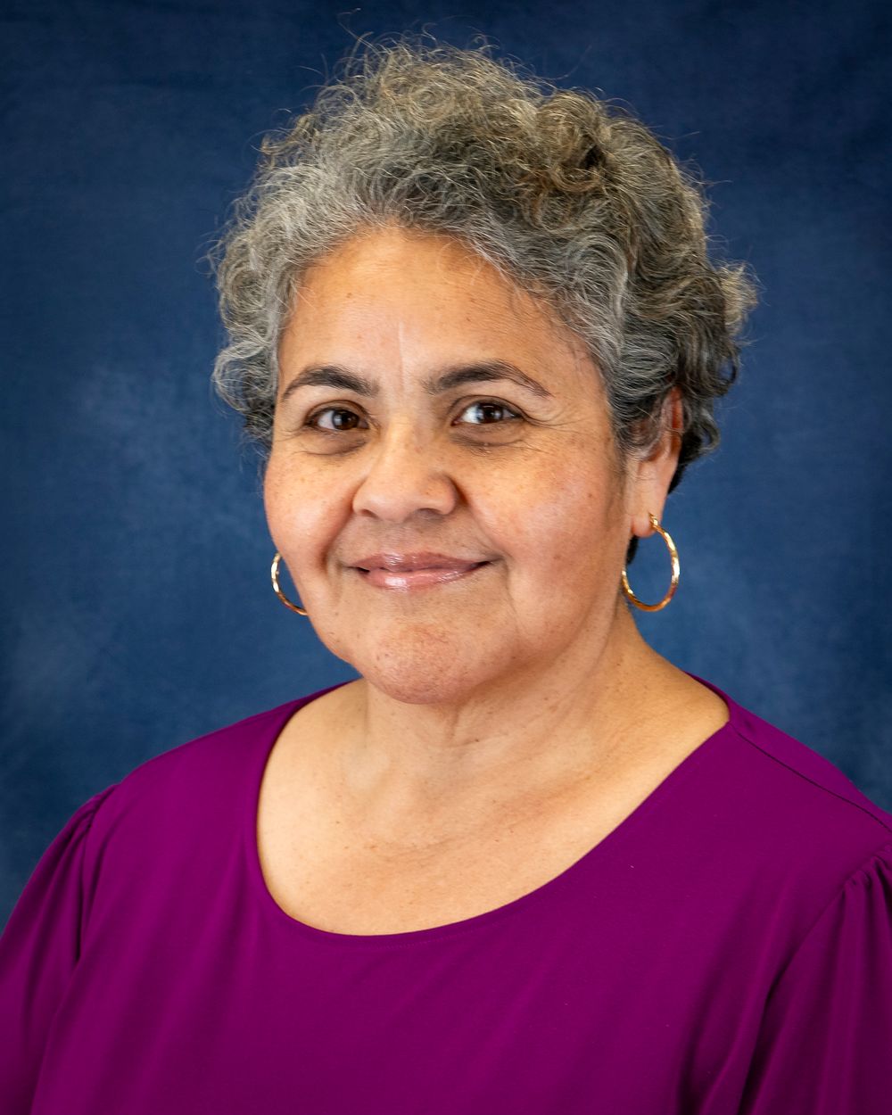Portrait of a person with short curly hair in a purple top on a blue backdrop.