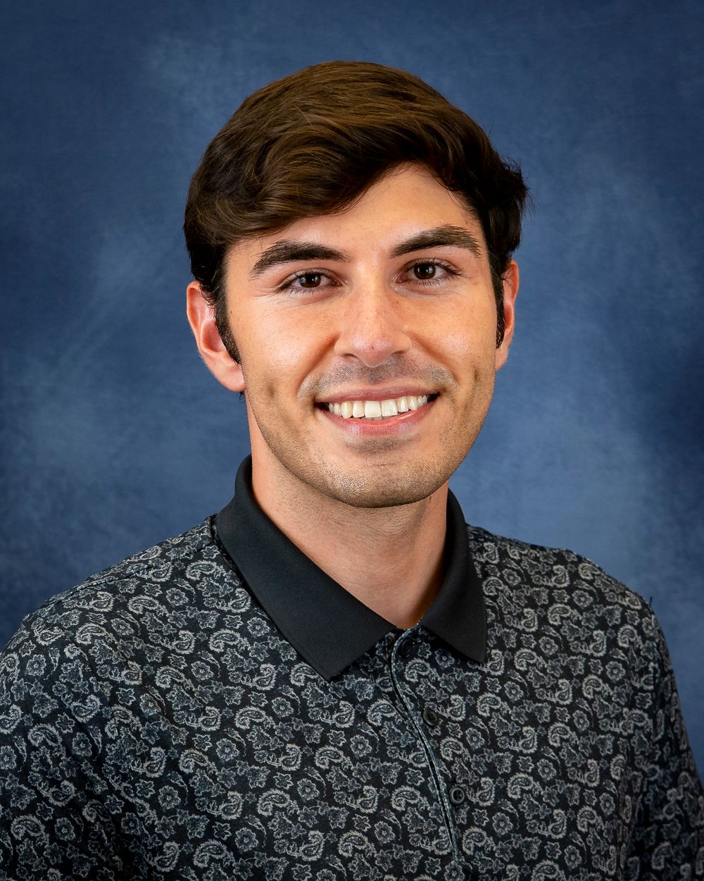Portrait of a person in a patterned dark polo shirt against a blue studio backdrop.
