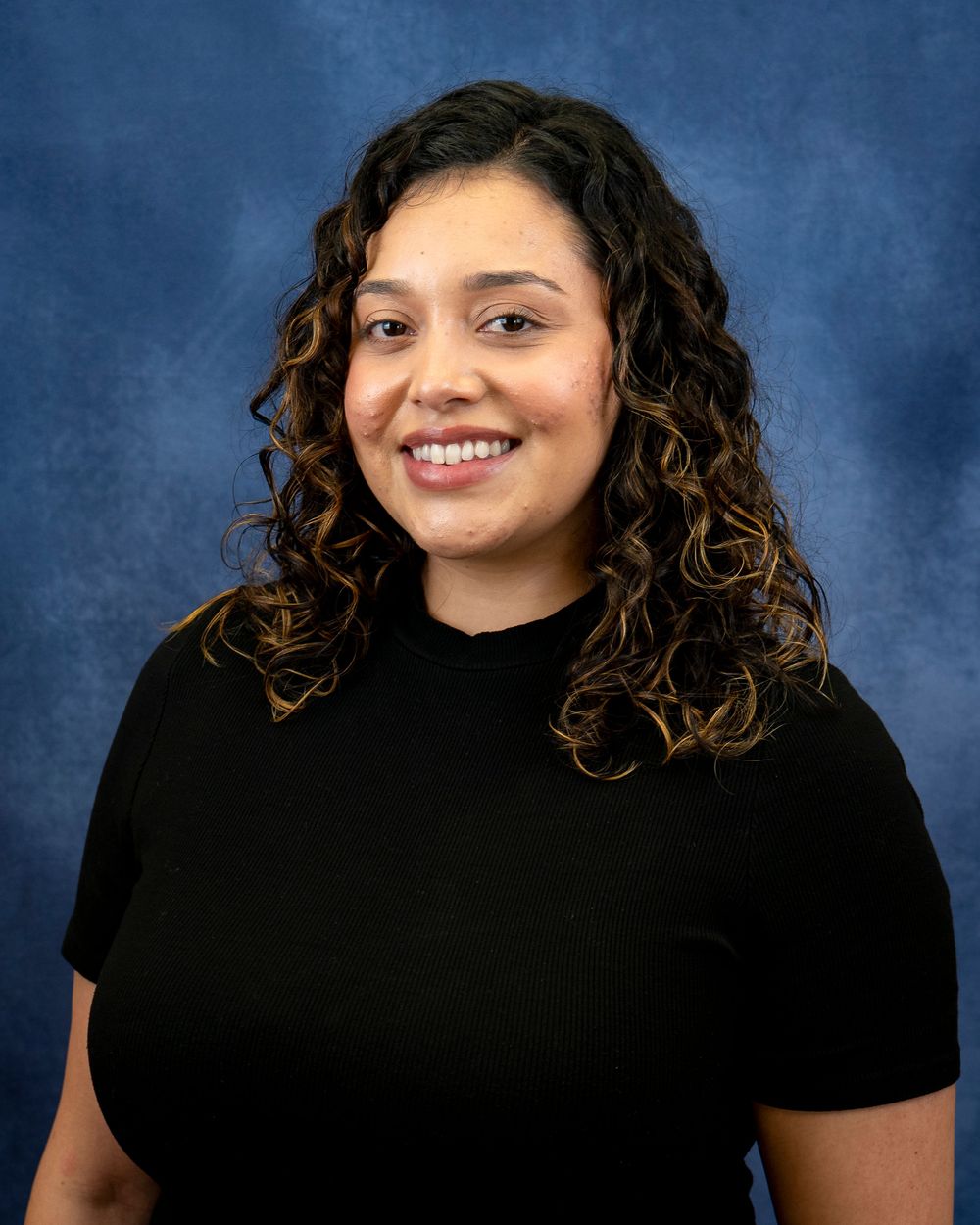 Portrait of a person with curly hair wearing a black top on a blue backdrop.
