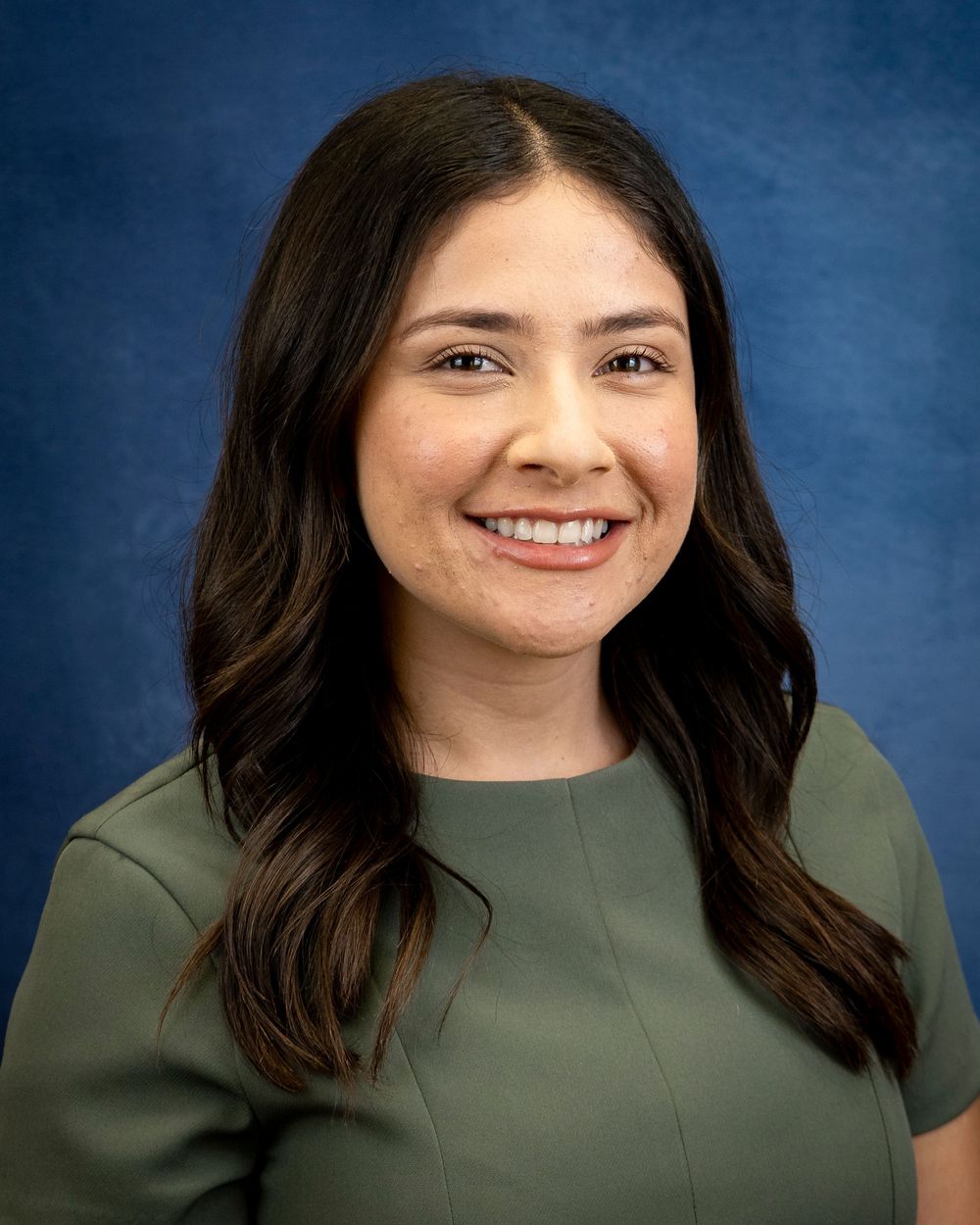 Portrait of a person with long dark hair wearing an olive green top on blue backdrop.