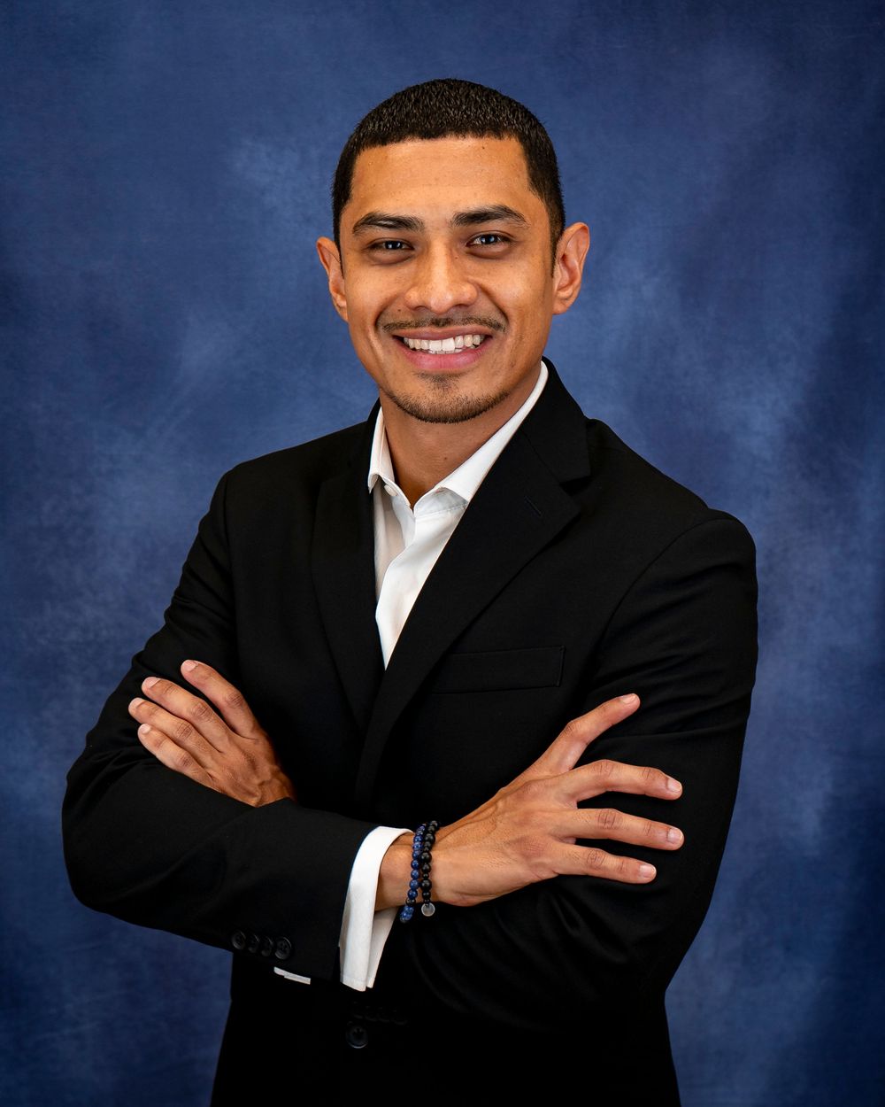 Smiling person in a black suit and white shirt posing with arms crossed on blue backdrop.
