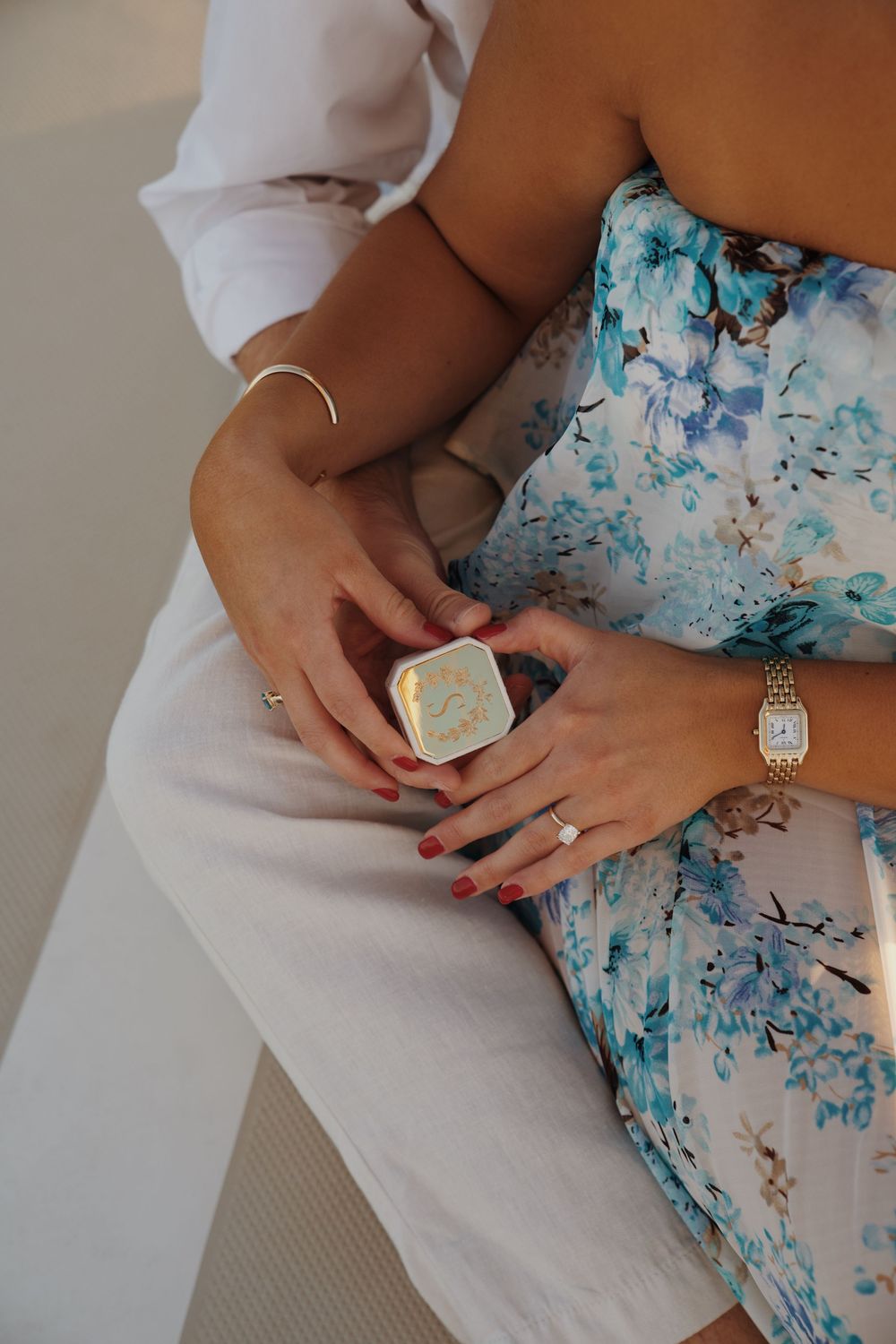 Close-up of hands holding a ring box while wearing a floral blue dress and white clothing.