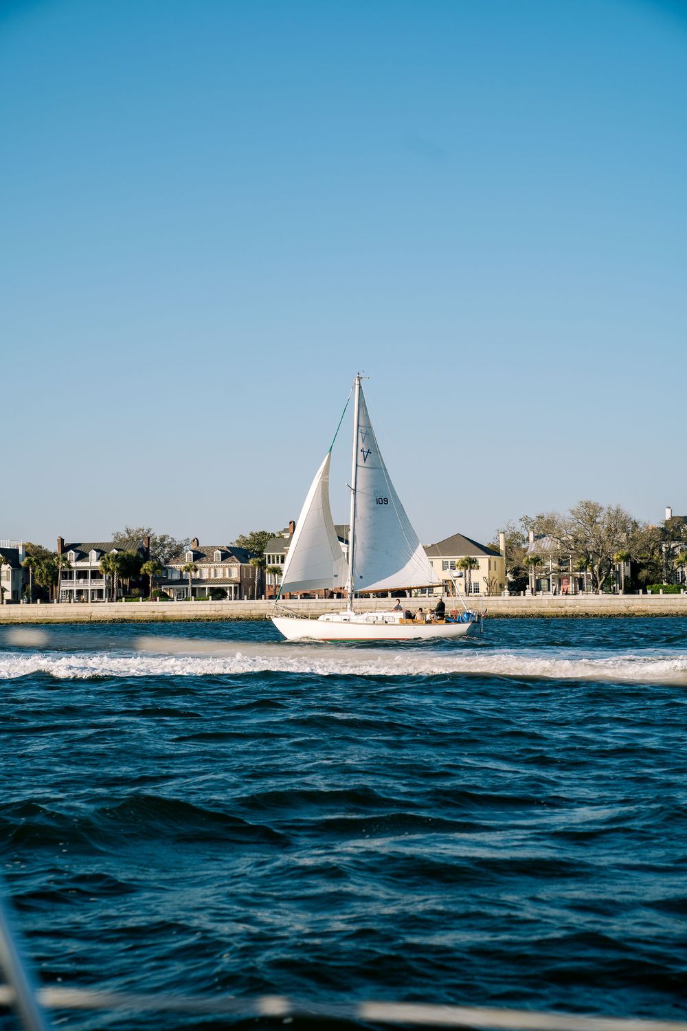 White sailboat cruising past beachfront homes on blue waters.