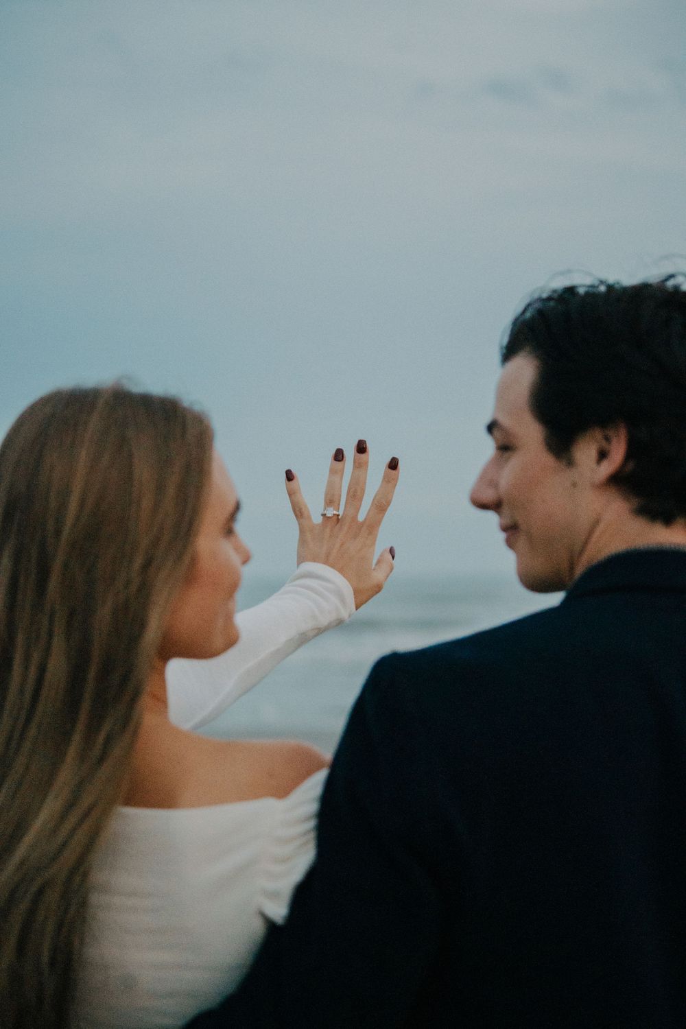 A couple shares an intimate moment against a cloudy sky backdrop as she reaches out to touch his face.