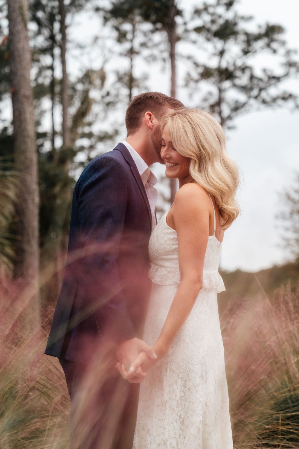 A romantic wedding couple embraces in a dreamy outdoor setting with tall pine trees and wispy grass.