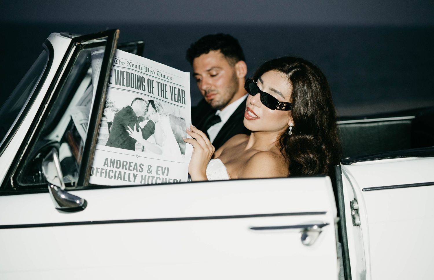 Editorial wedding shot—bride in sunglasses holds a “Wedding of the Year” paper in a classic car; groom in background.