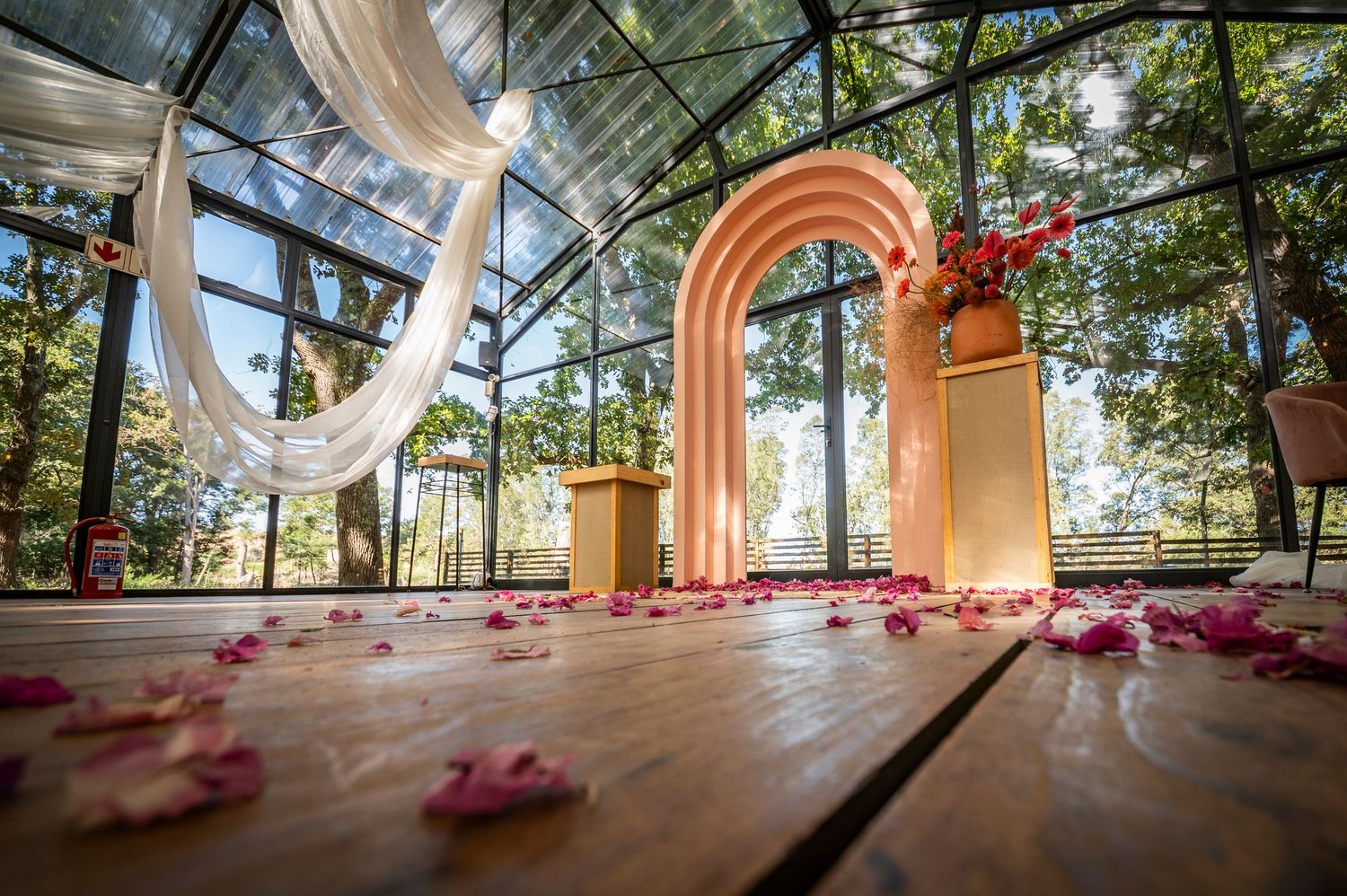 Wooden deck with pink rose petals leads to an elegant peach-colored archway in a glass-enclosed garden setting.