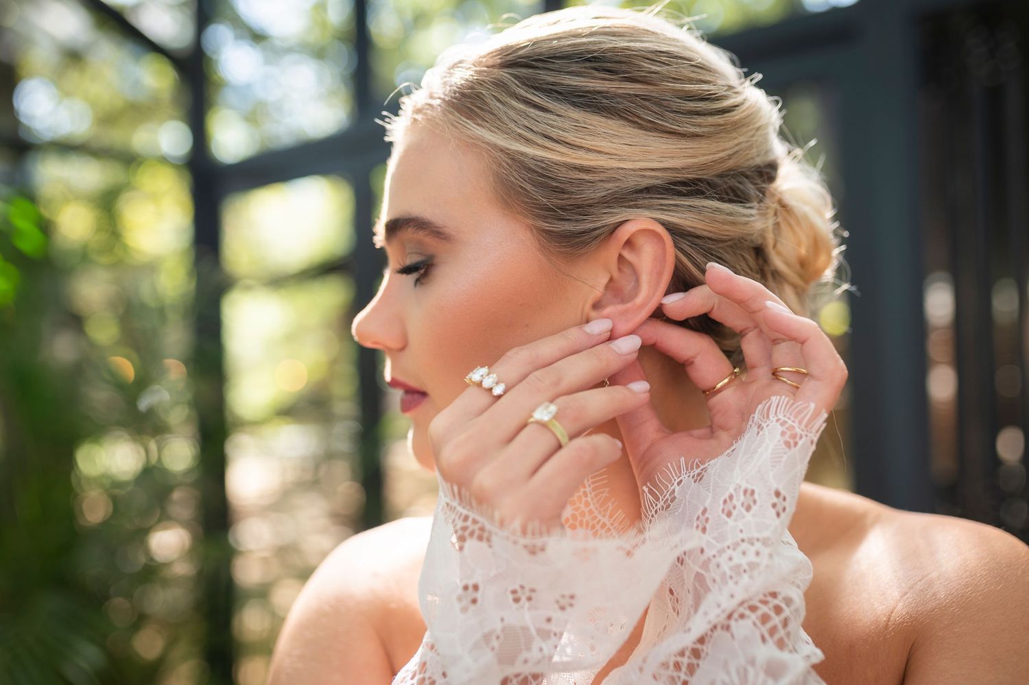 A bride in a delicate lace wedding dress getting ready in a sun-dappled forest setting.