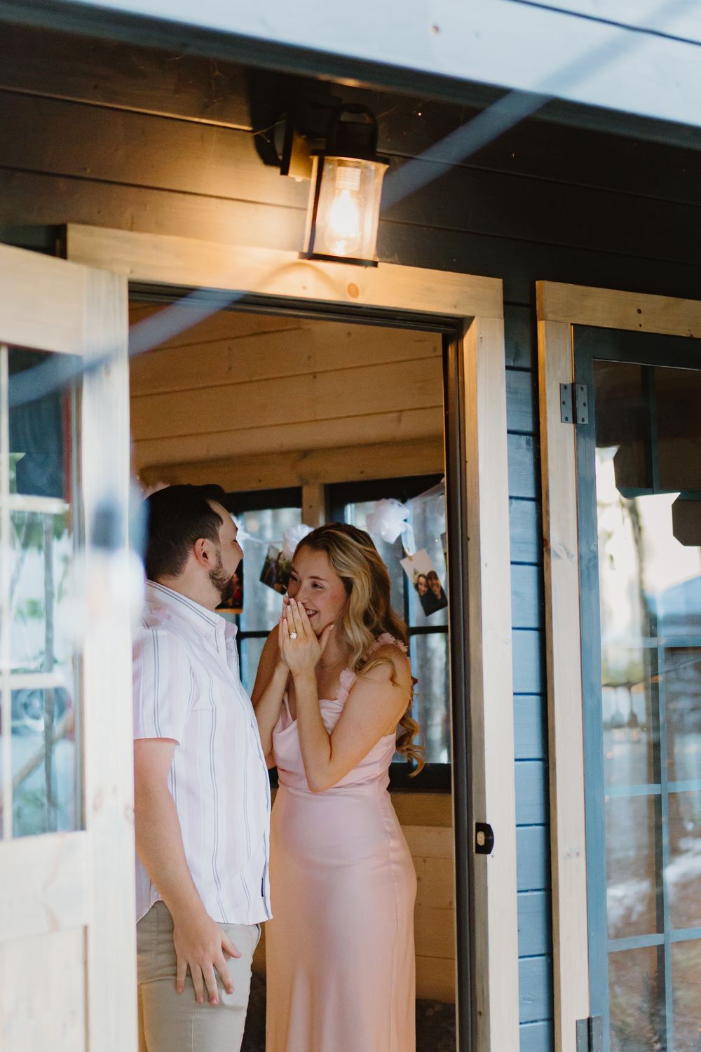 A romantic couple in white and pink attire share an intimate moment on a porch with rustic lighting.