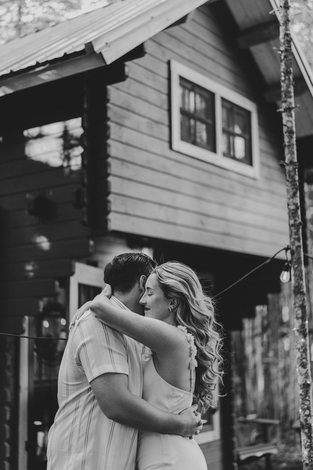 A romantic embrace and kiss between a couple in front of a rustic cabin with string lights, captured in black and white.
