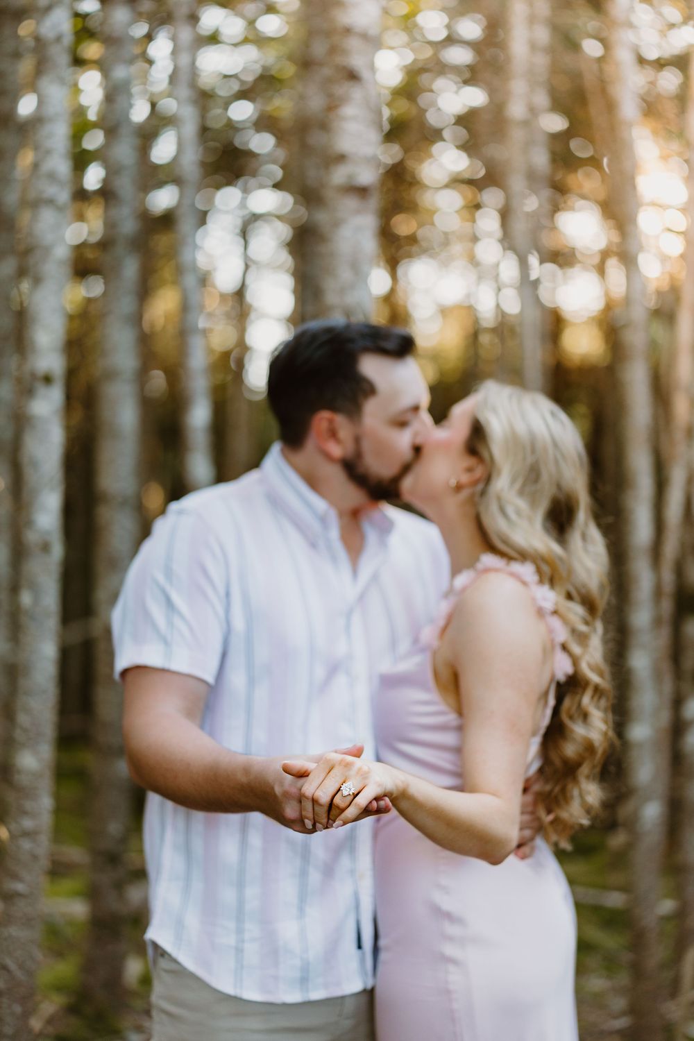 A romantic engagement portrait of a couple sharing a kiss in a sunlit forest with soft bokeh lighting effects in the background.
