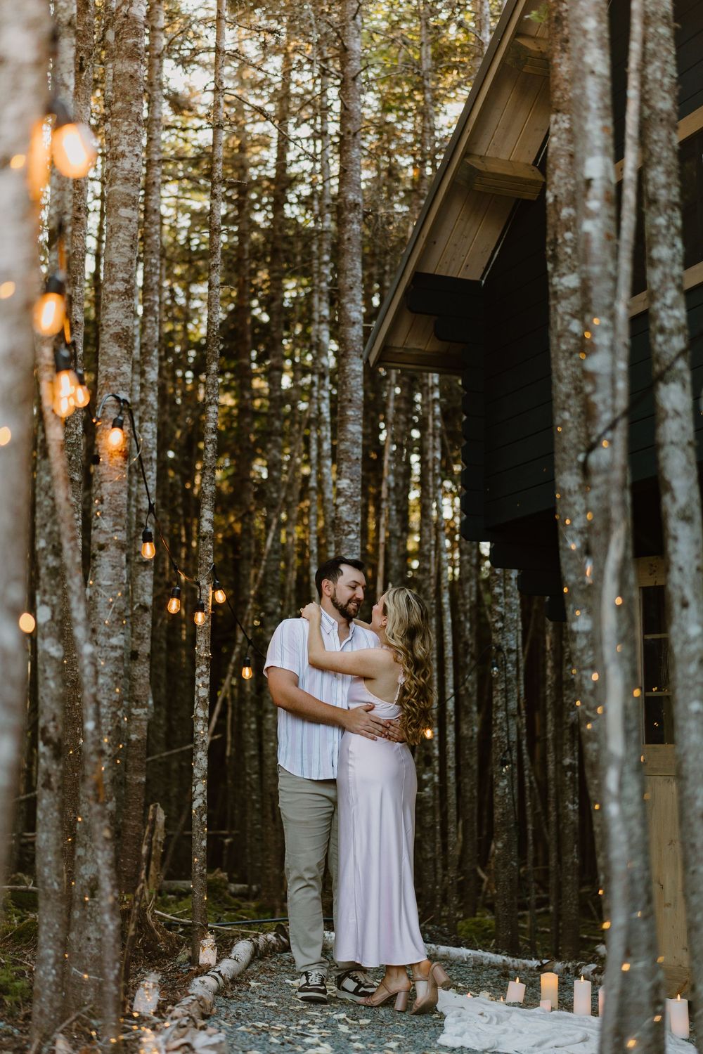 A couple embraces on a pathway lined with string lights and birch trees at dusk.