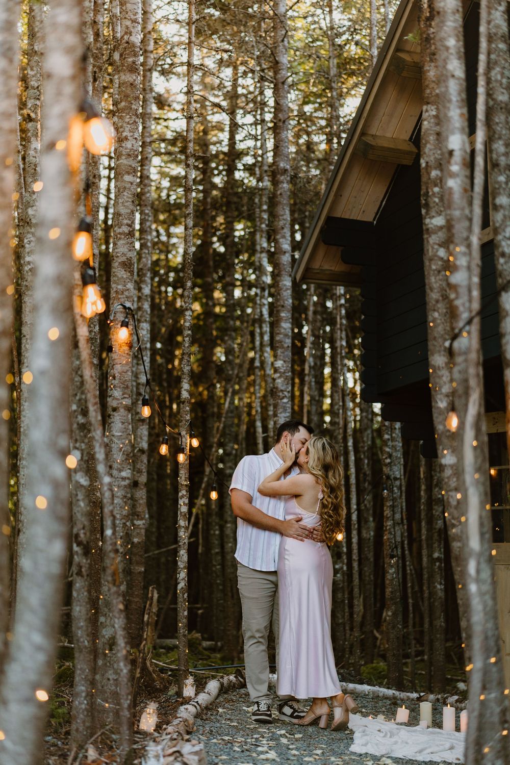 A romantic couple embraces in a forest setting with twinkling string lights and a wooden cabin at sunset.