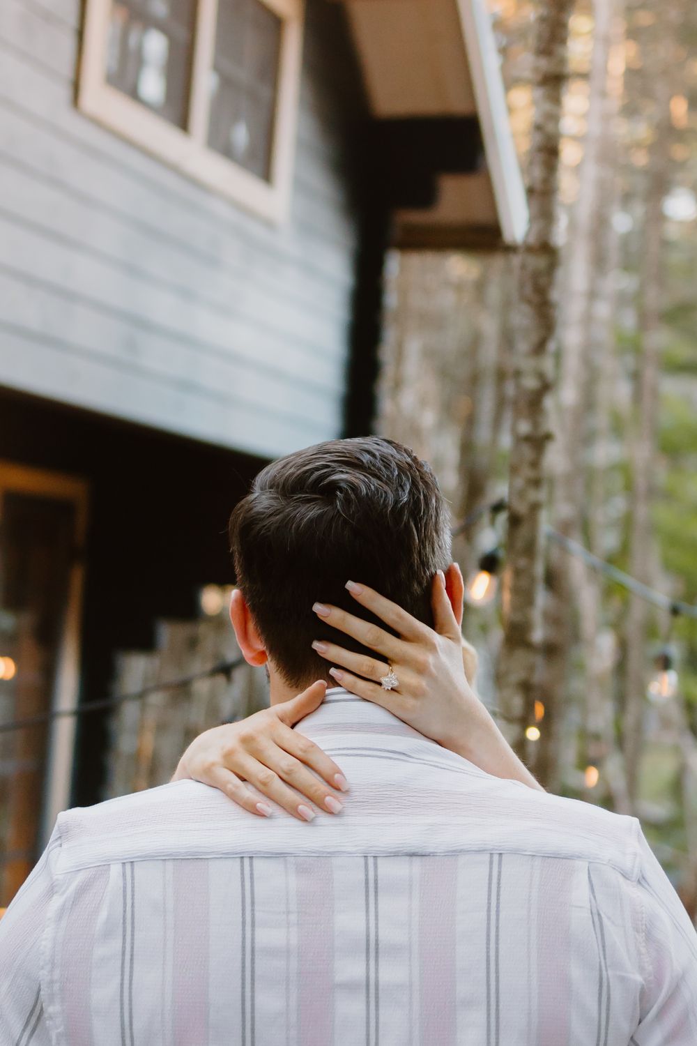 Back view of a person in a white striped shirt receiving a neck massage outdoors near a building with trees in background.