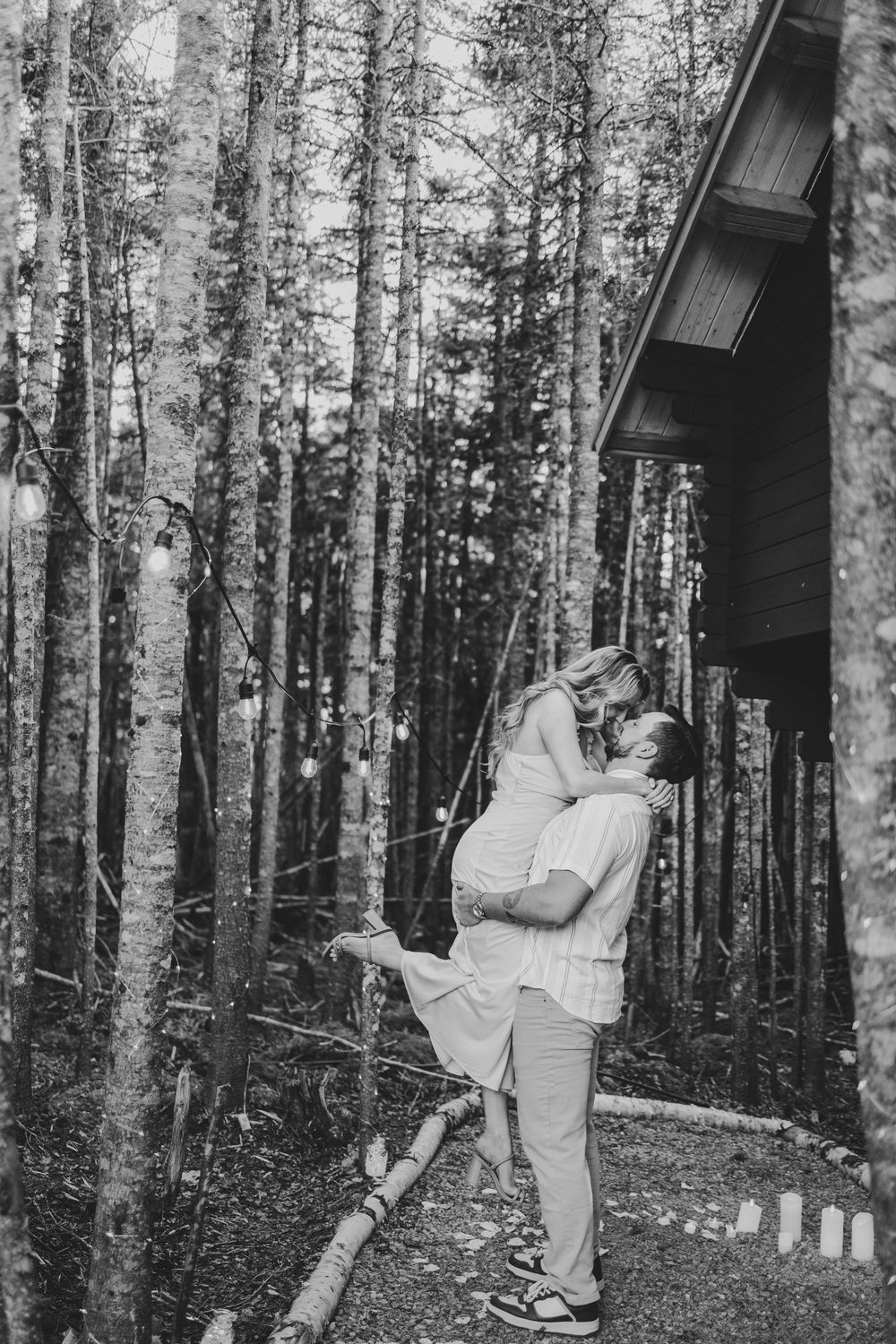 A couple in matching white outfits shares a romantic dance together in a forest setting near a rustic cabin.