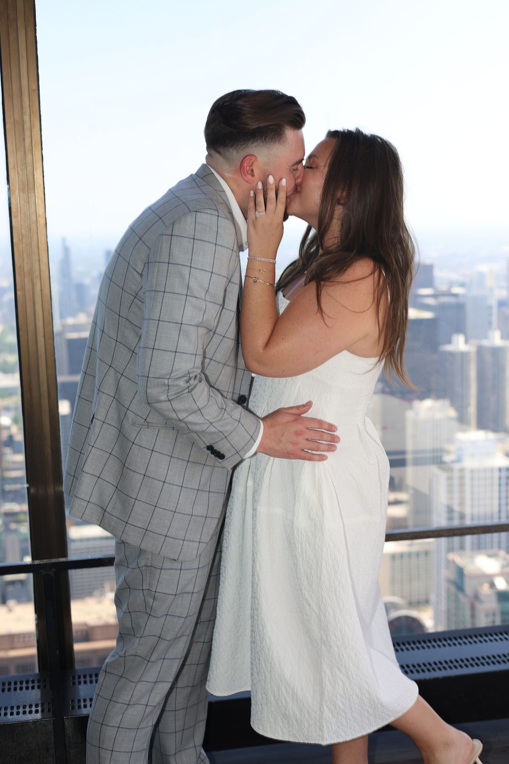 A couple shares a romantic kiss at a high-rise observation deck overlooking a cityscape.