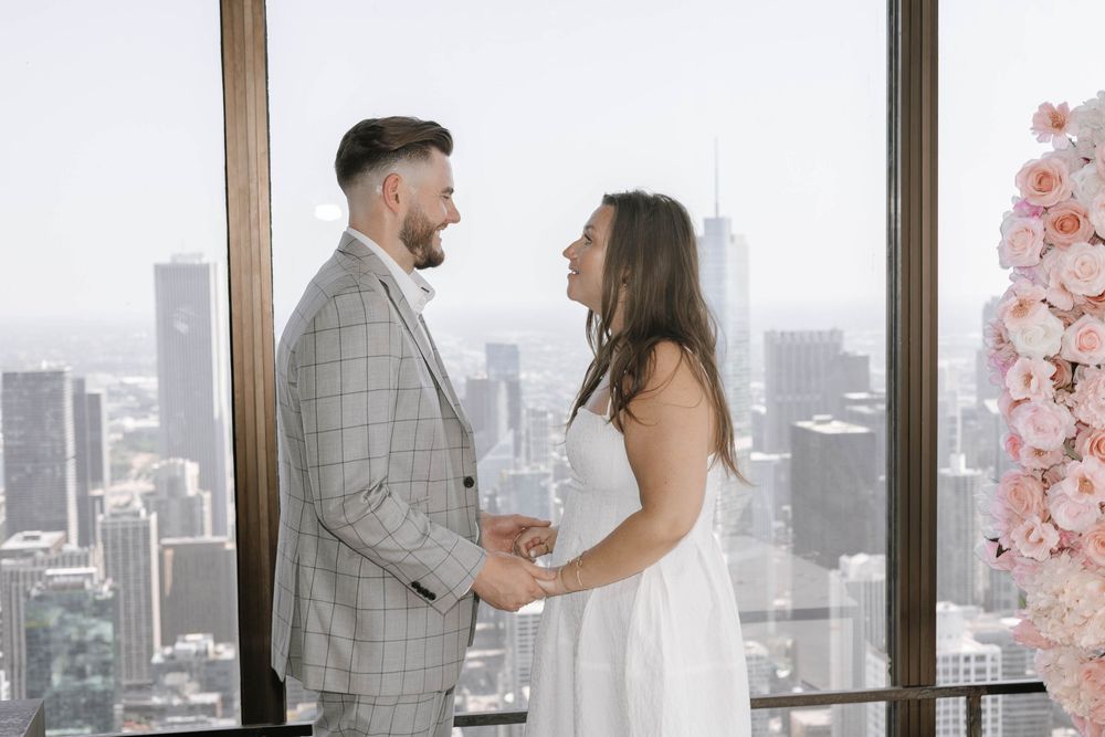 A couple shares an intimate moment in front of large windows overlooking a cityscape with pink cherry blossoms nearby.