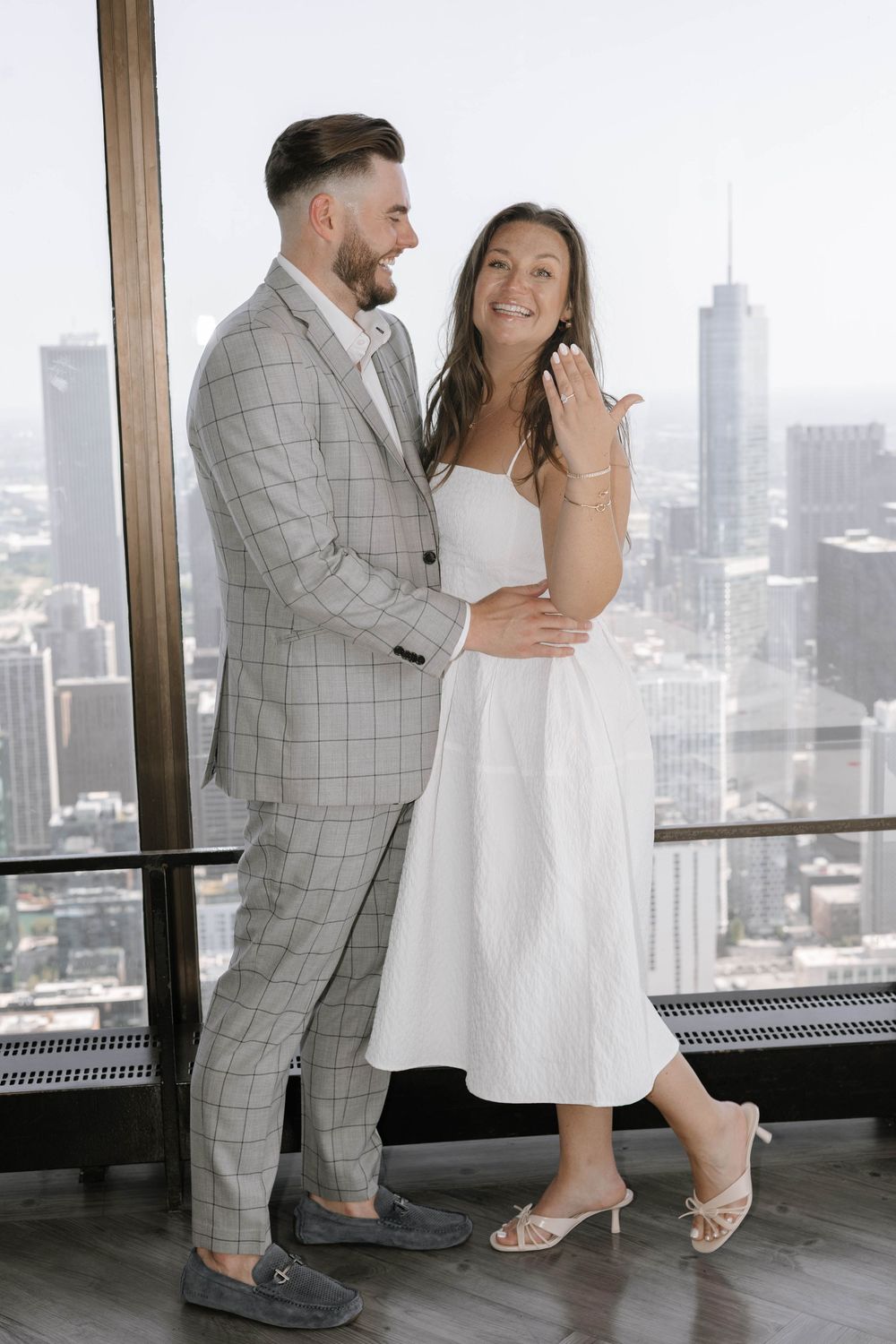 A couple shares an intimate moment in front of floor-to-ceiling windows overlooking a city skyline.