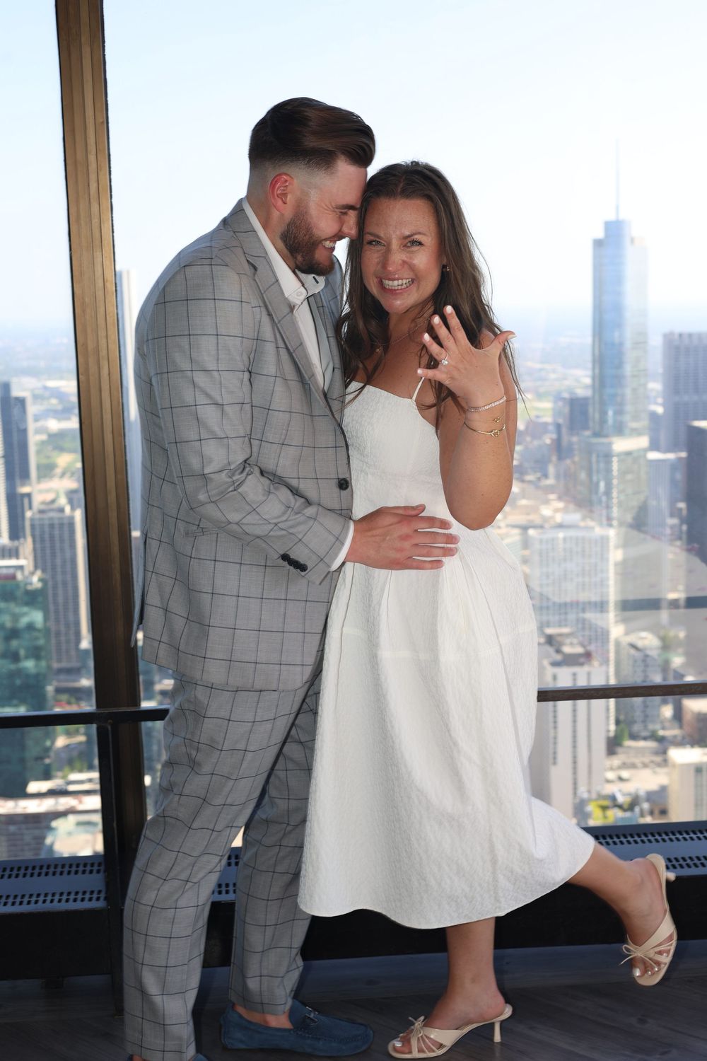 A newly engaged couple sharing an intimate moment at an observation deck overlooking a cityscape through glass windows.