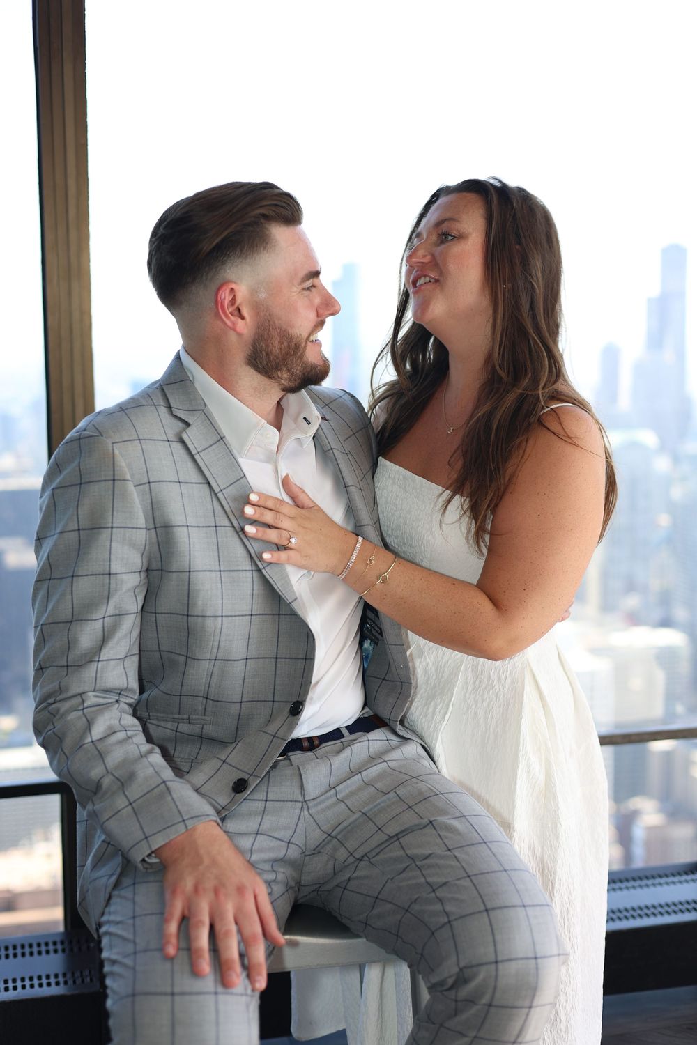 A loving couple shares a romantic moment in front of city skyline views, wearing elegant white and grey attire.