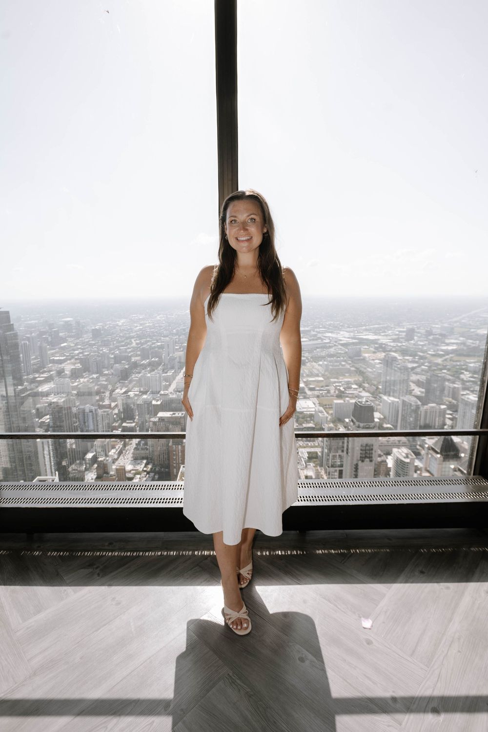 A person in a white summer dress stands at an observation deck overlooking a city skyline view.