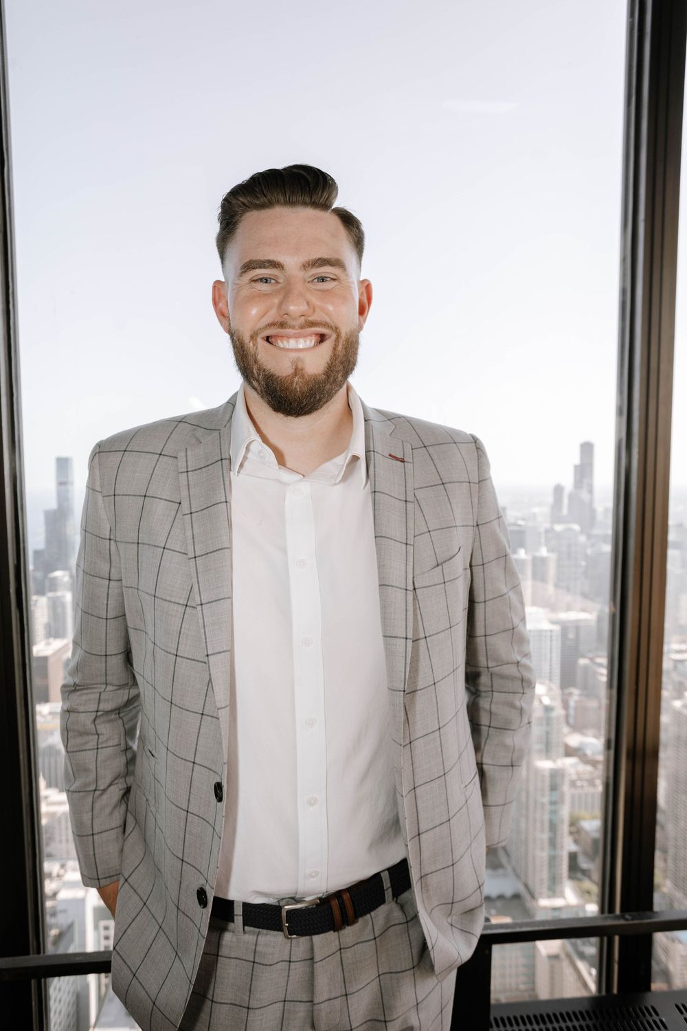 Professional wearing a gray plaid blazer and white shirt stands smiling in front of large office windows with city skyline view.