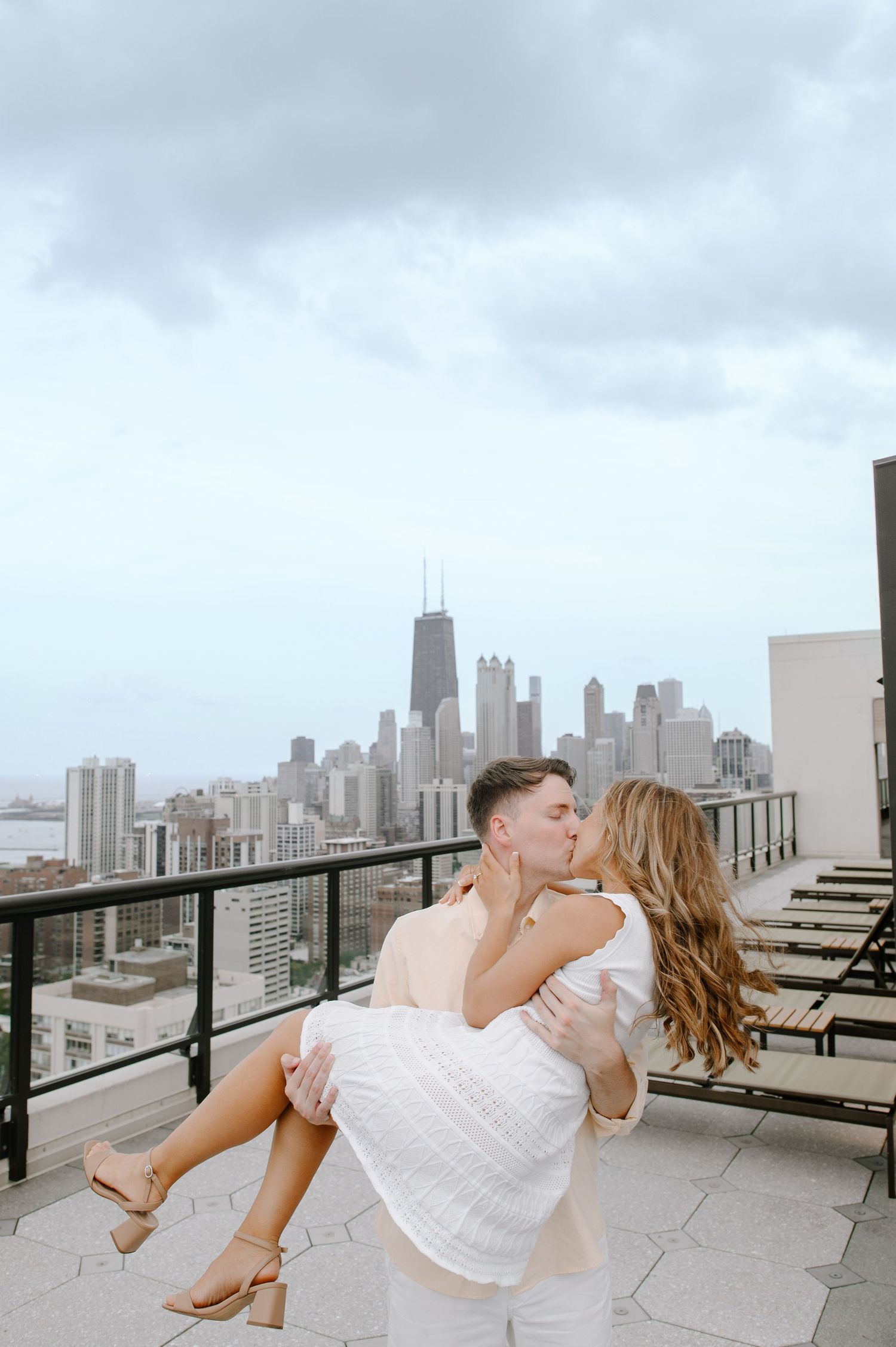 A romantic couple shares an intimate moment on a rooftop terrace overlooking the Chicago skyline on a cloudy day.