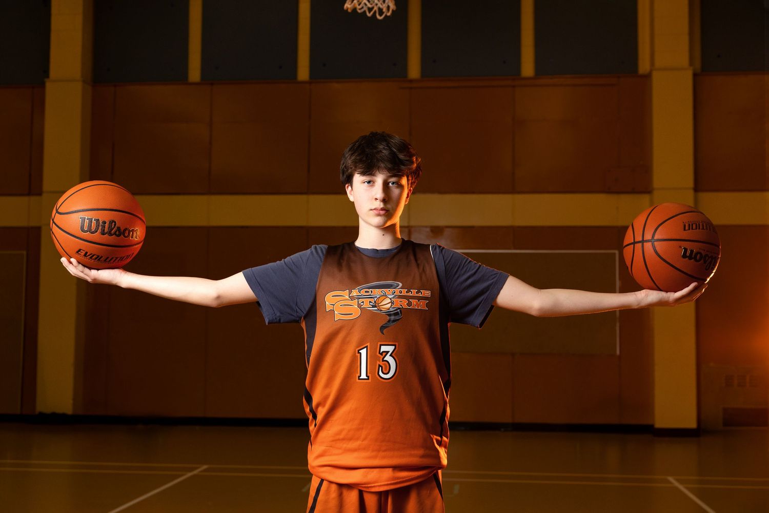 Basketball player wearing orange jersey number 13 holds two basketballs out to sides in indoor gymnasium.