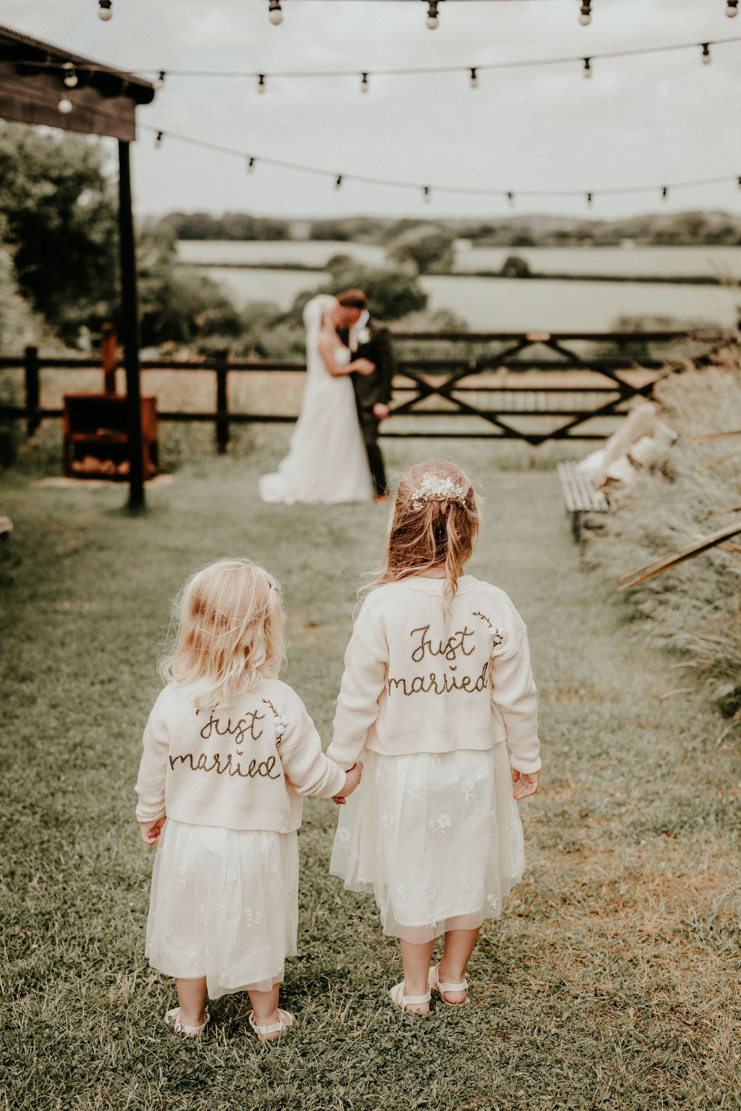 Two matching denim jackets with 'Just Married' text walk through a rustic outdoor setting with string lights overhead.