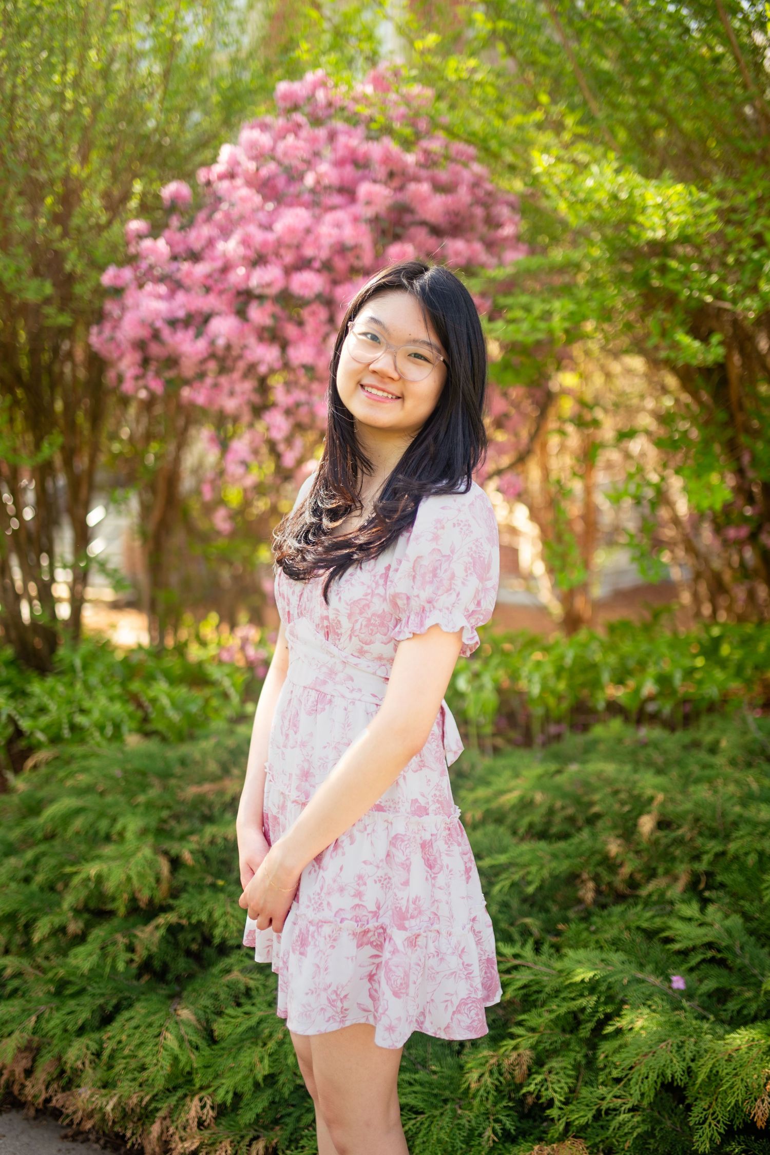 A person in a pink floral dress stands in a garden with blooming azaleas and ferns in the background.