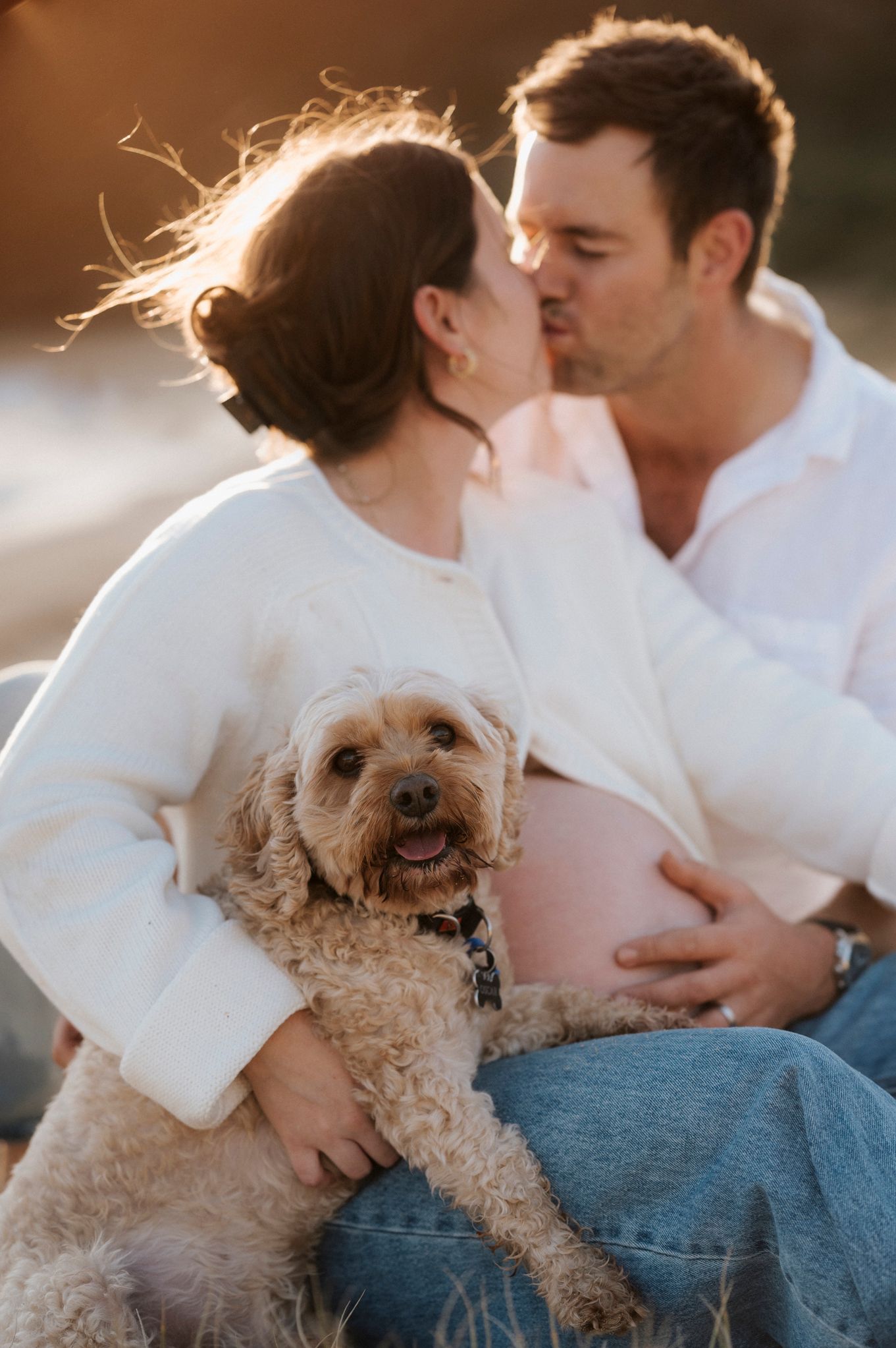 A couple shares an intimate moment outdoors with their small curly-haired dog during a maternity photo session.