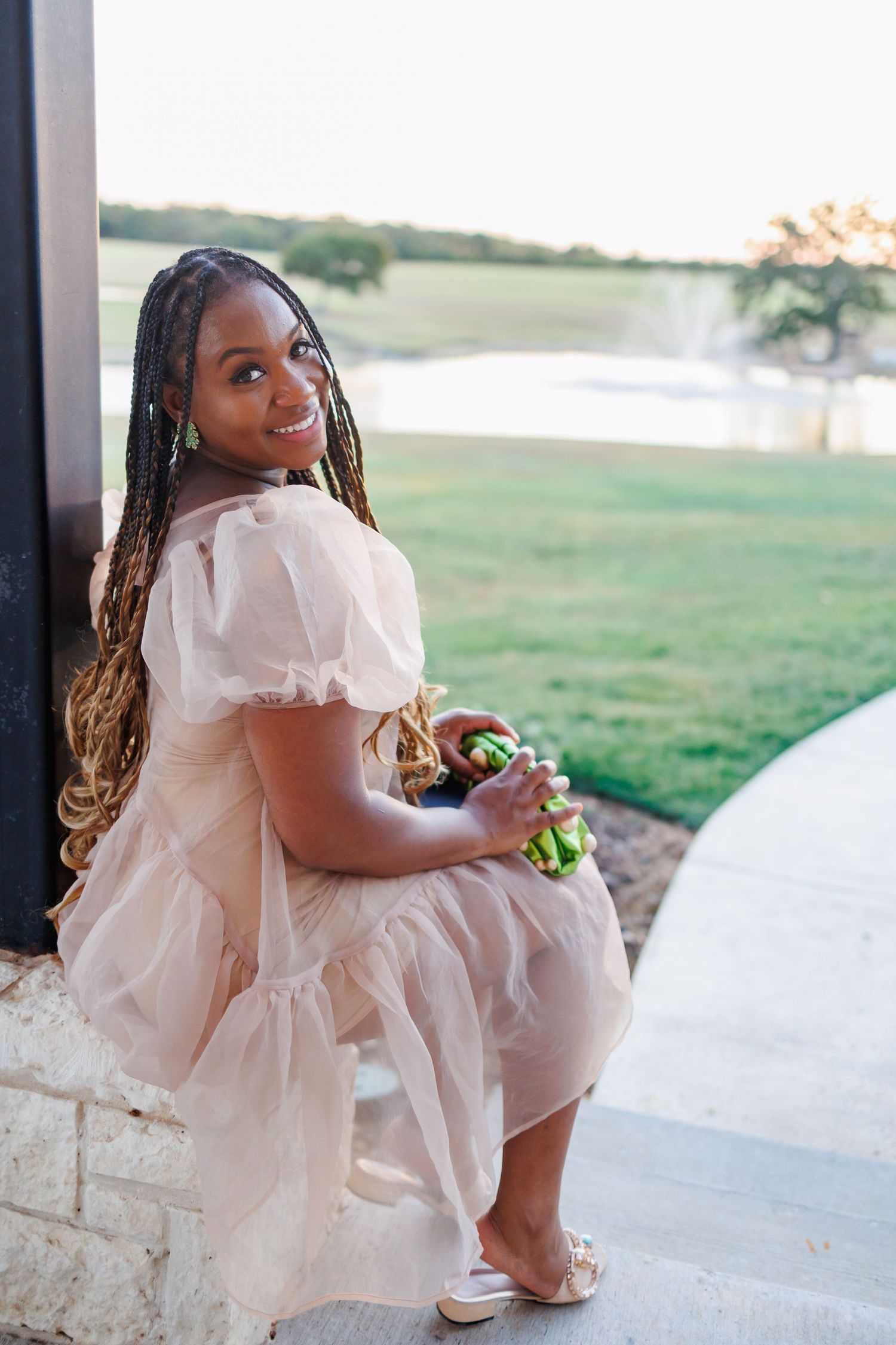 A woman in a flowing blush pink dress sits on a stone ledge at sunset overlooking the lawn at Bella Cavalli