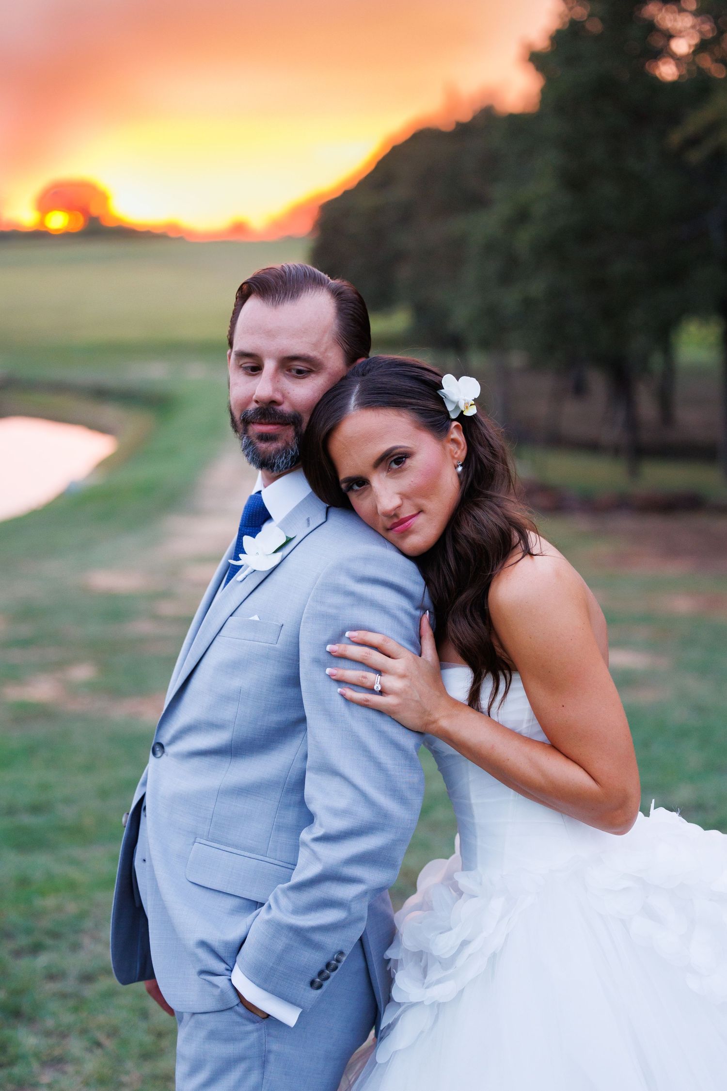 A bride and groom in wedding attire embrace against a sunset backdrop at Bella Cavalli with orange sky and trees in background.