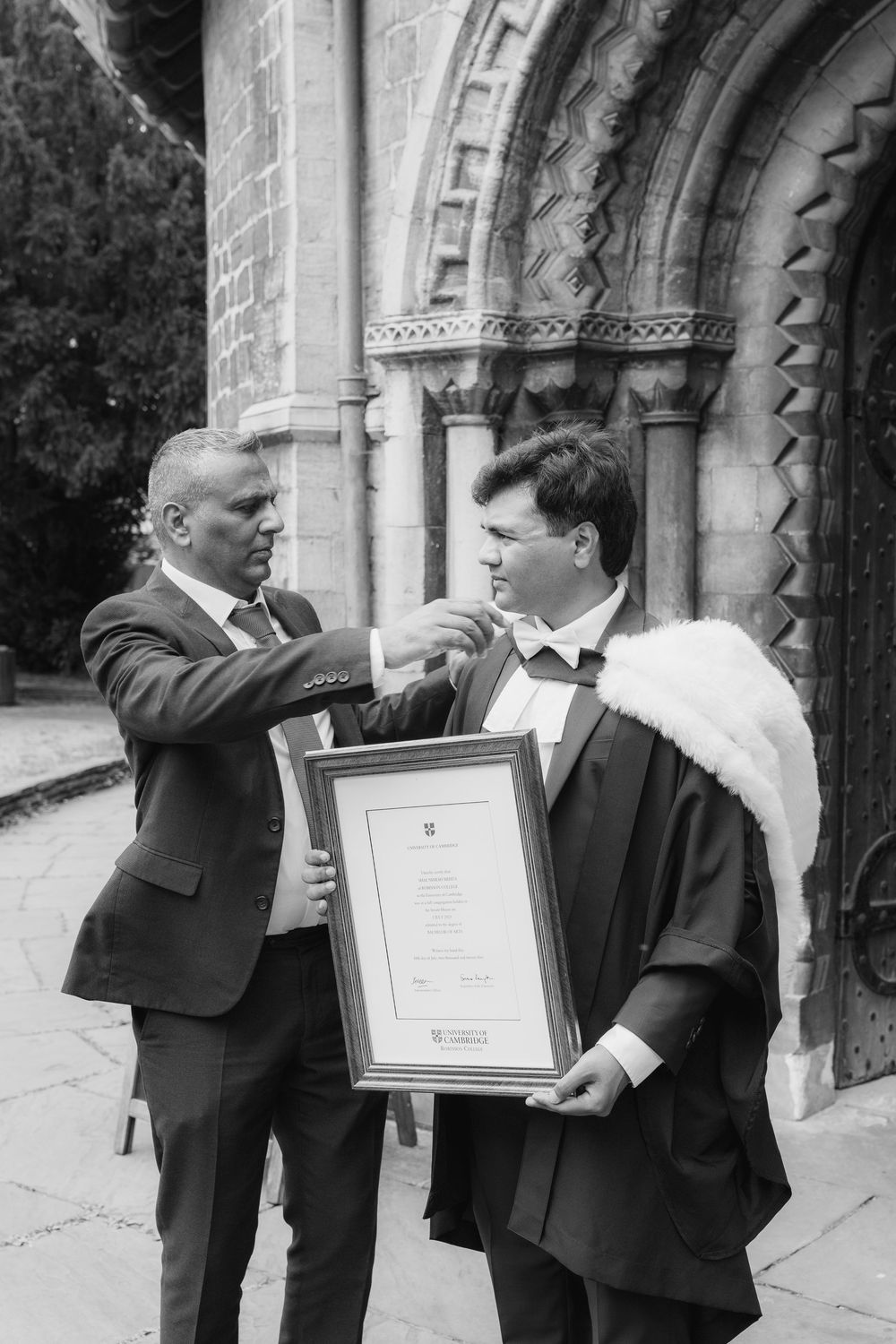 Two men in suits stand outside a stone building examining framed documents together.