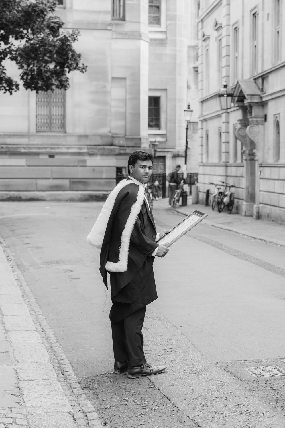 A vintage black and white photo of a college student in academic regalia standing on a sidewalk near classic buildings.