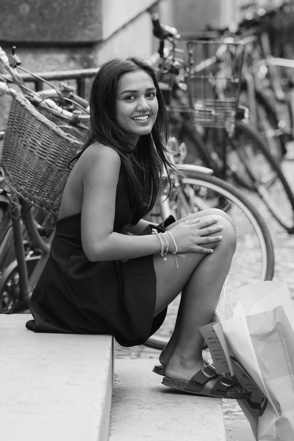 A person in a black tank top sits on the ground next to bicycles while holding shopping bags and smiling.