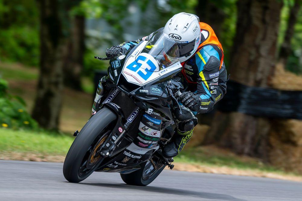 Motorcycle racer number 63 leaning into a curve on a black sport bike during a road race surrounded by green trees.