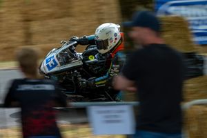 Motorcycle racer wearing white helmet speeds past hay bales on track while spectator watches in blurred foreground.