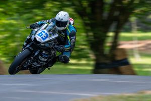Motorcycle racer number 13 leaning into a curve at high speed on a tree-lined racing circuit.