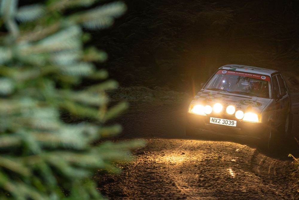Rally car speeding through a dark forest road with bright headlights illuminating the night.