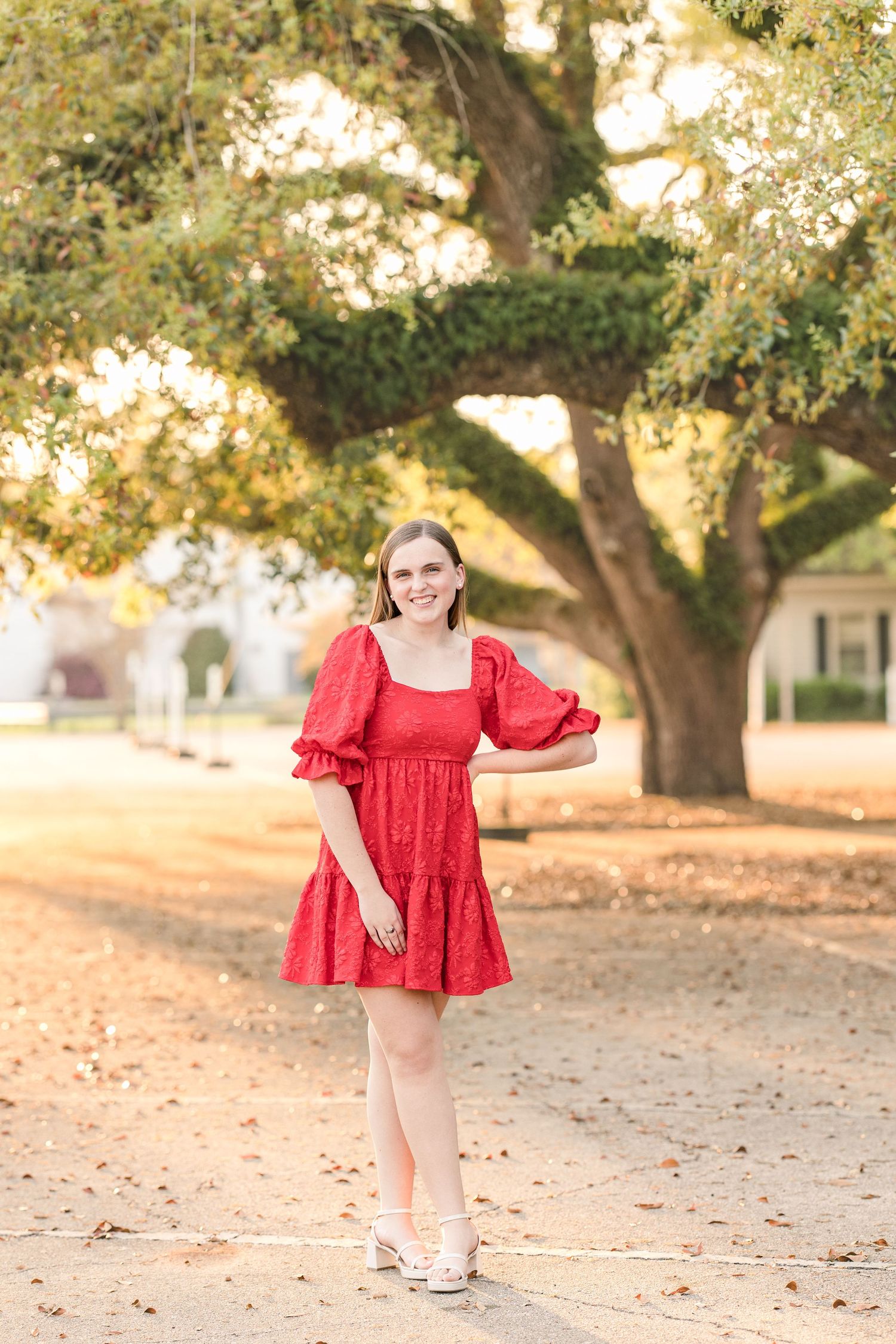 Someone in a red ruffled dress poses outdoors under a large oak tree on a fall day with autumn leaves scattered on the ground.