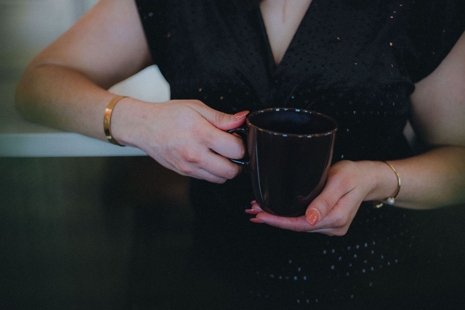 Person in black attire stands against dark wall while holding a white cup thoughtfully.