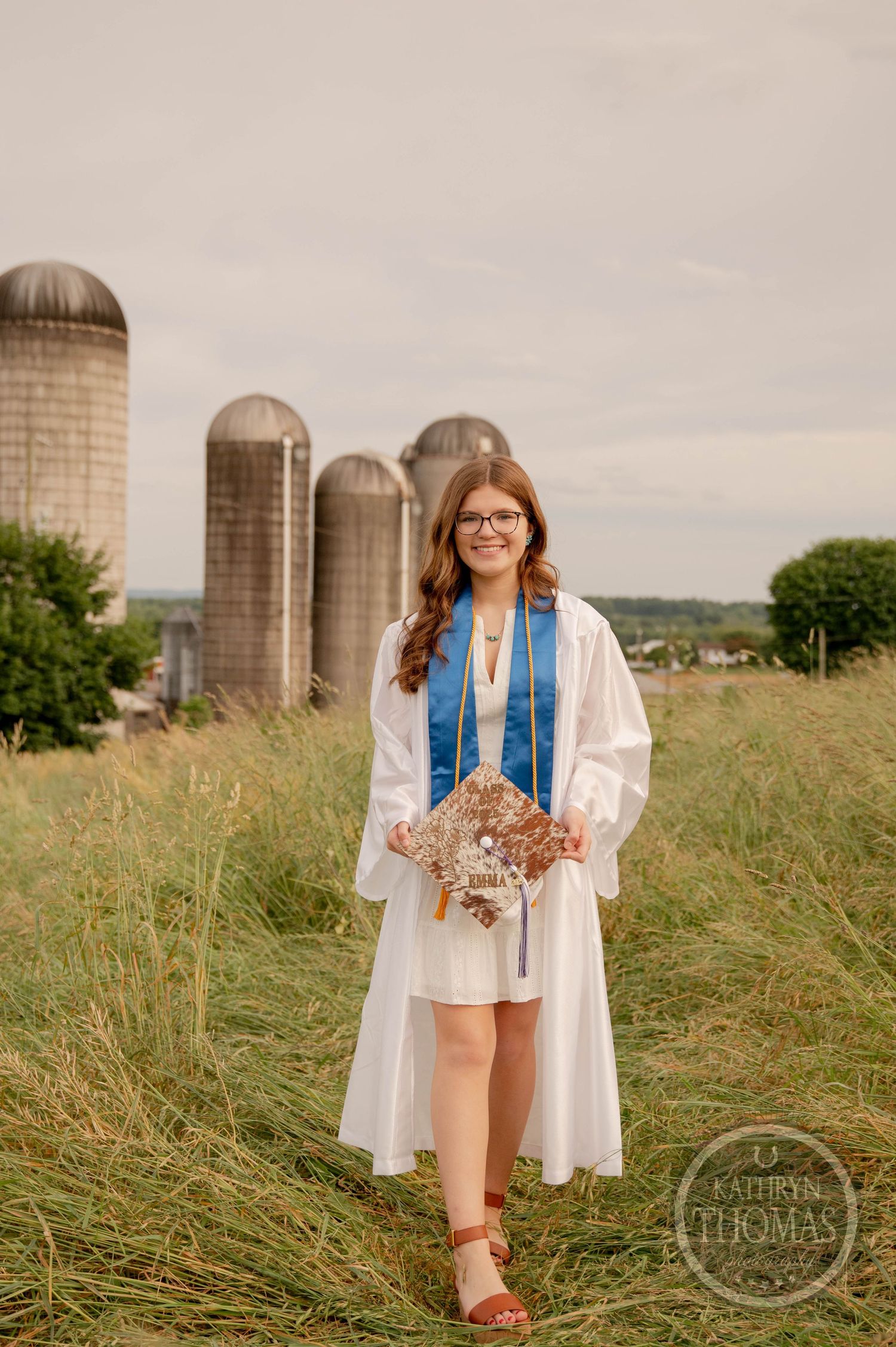 A graduation photo taken on a rural farm with tall silos in the background on a cloudy summer day.