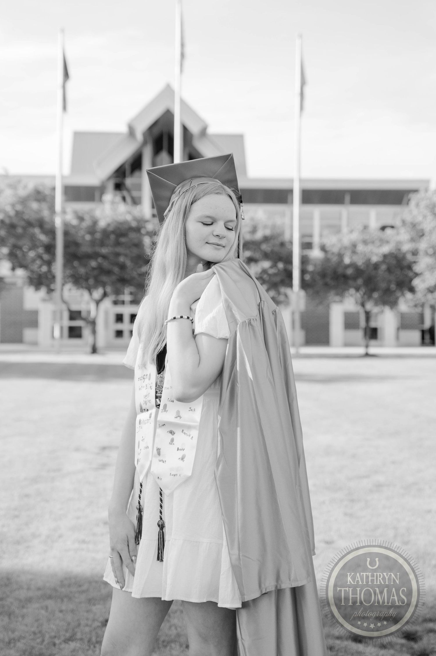 Graduate poses in cap and gown for graduation photos on campus grounds in black and white.