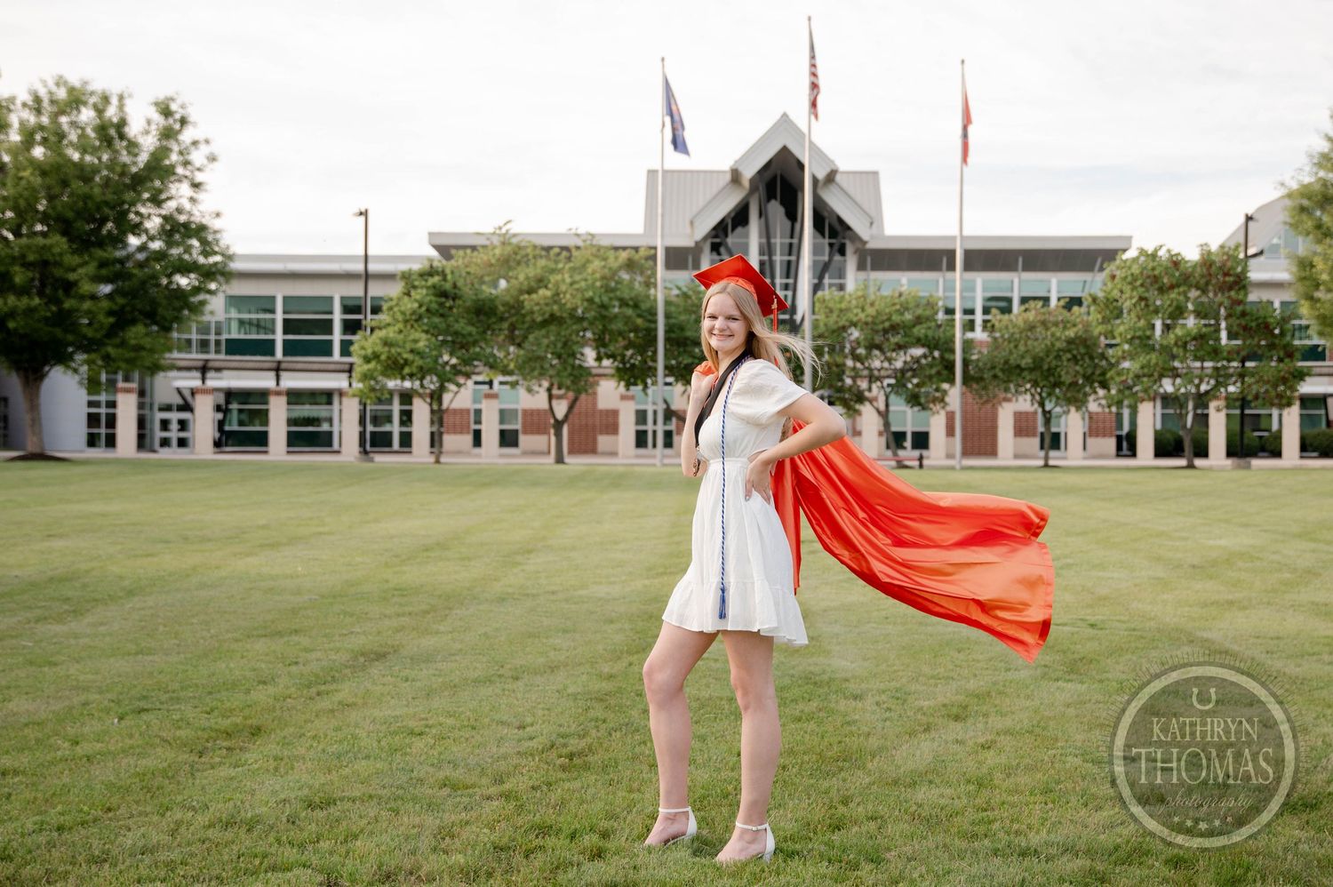 A person in a graduation gown stands on a campus lawn with a flowing red cape caught in the wind.