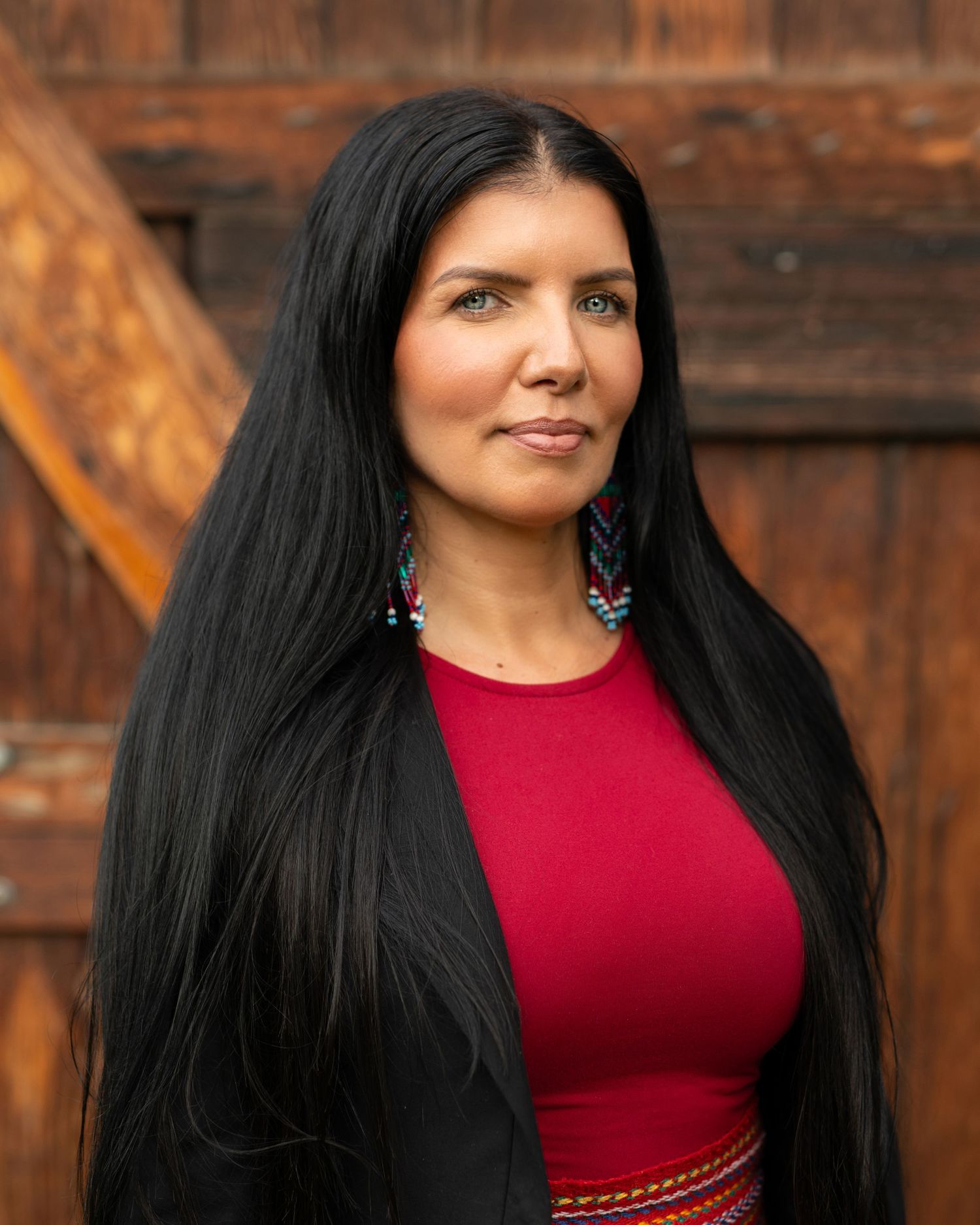 Woman with long dark hair wearing a red top and black blazer, standing in front of a wooden backdrop.